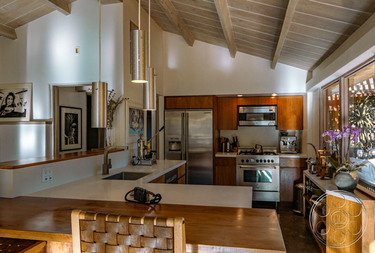 Pad 90210 - architectural style: Mid-Century Modern, minimalist design with clean lines, wood cabinetry with natural finish, pendant lighting over kitchen island, open concept layout with natural light, stainless steel appliances in modern kitchen, interior kitchen with exposed beam ceiling, concrete countertop with integrated sink, tile flooring with natural stone finish, built-in open shelving in kitchen, framed artwork as wall decor, large windows for outdoor view, wooden table with woven seat accents, neutral color palette with earth tones, kitchen area with indoor plants as décor