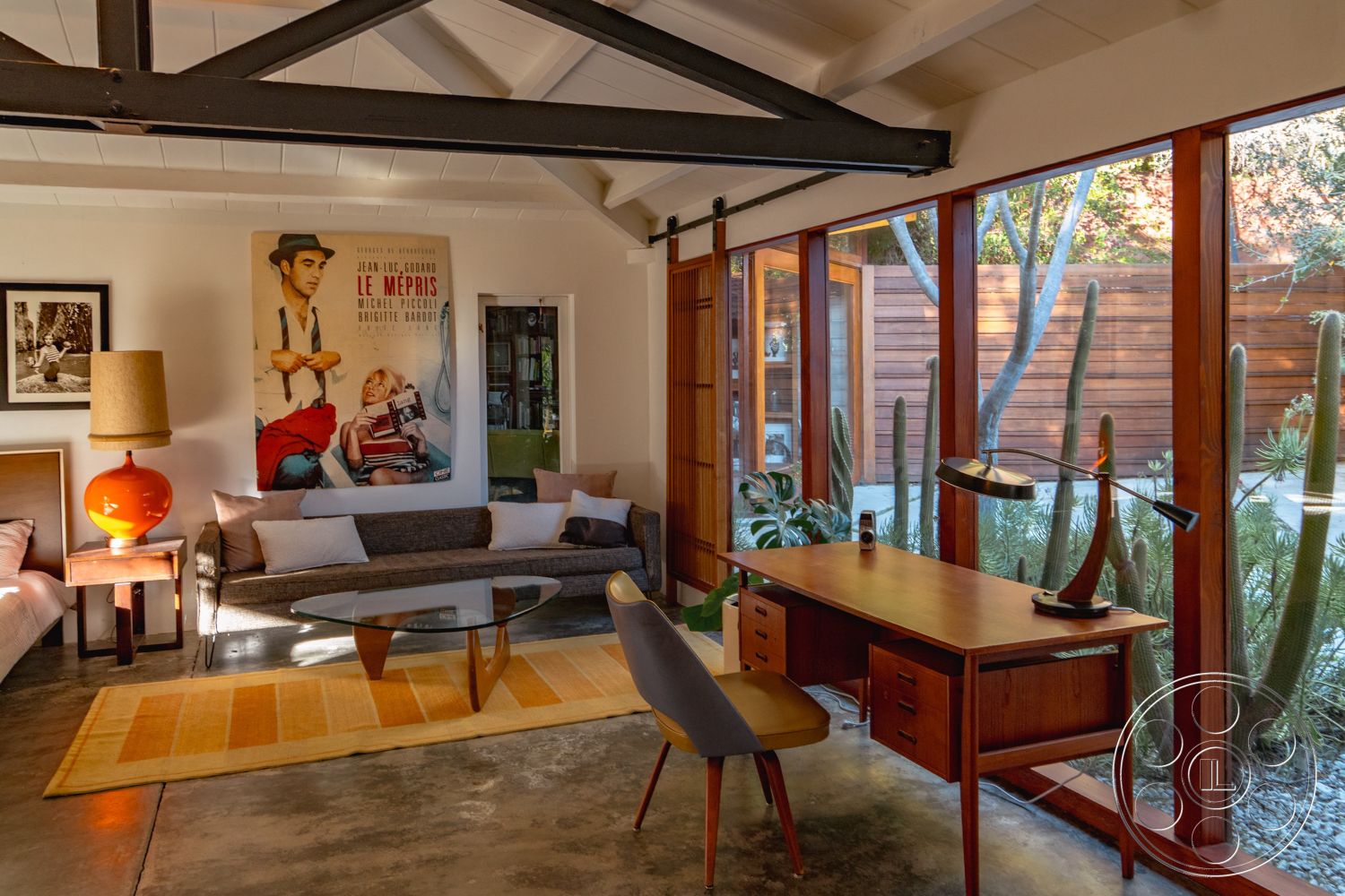 Pad 90210 - neutral color palette, bright natural lighting, contemporary armchair, yellow area rug, exposed wood ceiling beams, floor-to-ceiling glass windows, interior concrete flooring, minimalist decorative elements, modern glass coffee table, wooden desk with drawers, mid-century modern sofa, orange ceramic table lamp, wooden sliding door system, natural backdrop with cacti, vintage movie poster artwork, upholstered fabric sofa, saltillo tile wall