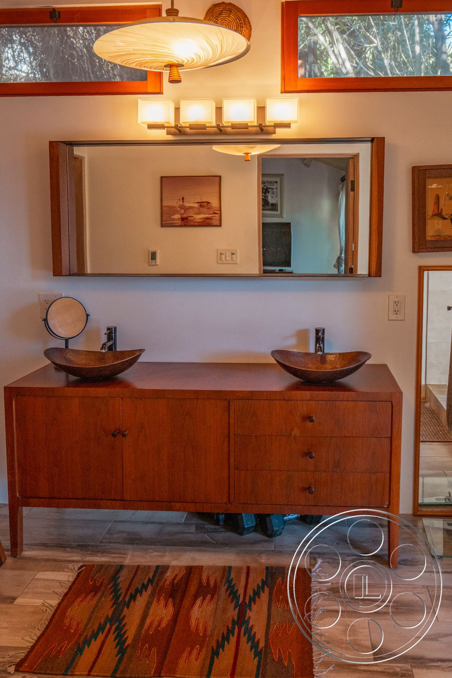 Pad 90210 - textured plaster walls, warm wood accents, exposed wood ceiling beams, tile bathroom flooring, large wall-mounted mirror, interior bathroom with wooden vanity, stone sinks on wooden vanity, mid-century modern lighting fixture, natural light from recessed windows, colorful woven area rug, minimalist cabinetry with wood finish, clean lines with modern fixtures, brightly lit bathroom vanity area, open concept design with adjacent rooms