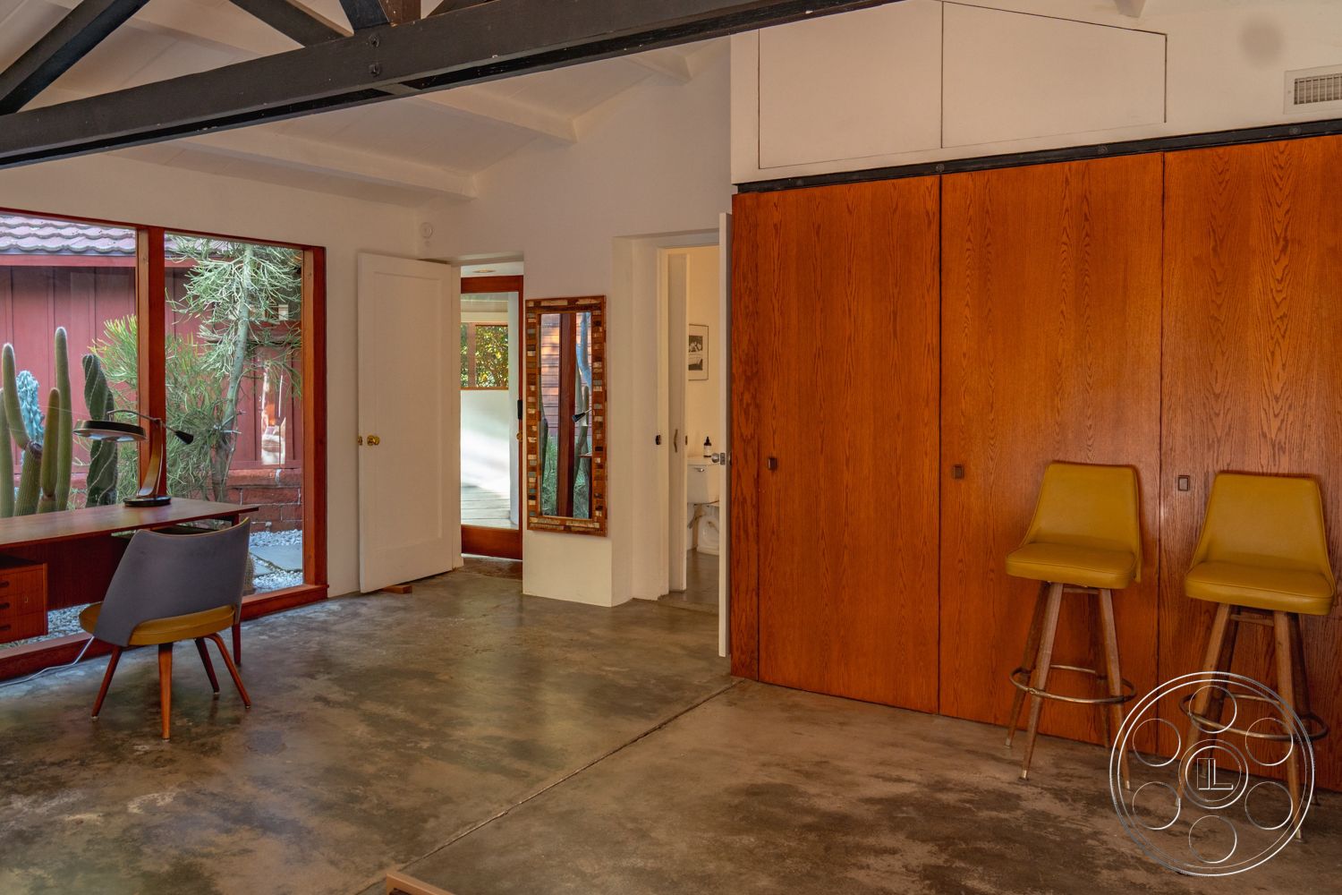 Pad 90210 - neutral color palette, large glass windows, interior garden view, indoor plant decor, open interior layout, natural light abundance, exposed concrete flooring, mid-century modern aesthetic, functional home office space, wood panel wall cabinets, steel beams ceiling support, minimalist workspace setup, modern desk and chair, compact bathroom access, sleek bar stools with wooden legs