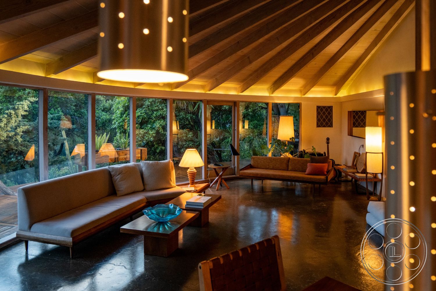 Pad 90210 - wooden coffee table, large floor-to-ceiling windows, wood accent elements, cozy lounge area, textured wall treatments, ambient lighting fixtures, exposed wood ceiling beams, natural light-filled space, open floor plan layout, minimalist furniture design, indoor plants accents, mid-century modern interior, secluded garden view, polished concrete living room floor, soft upholstered sectional sofa, decorative glass bowl centerpiece