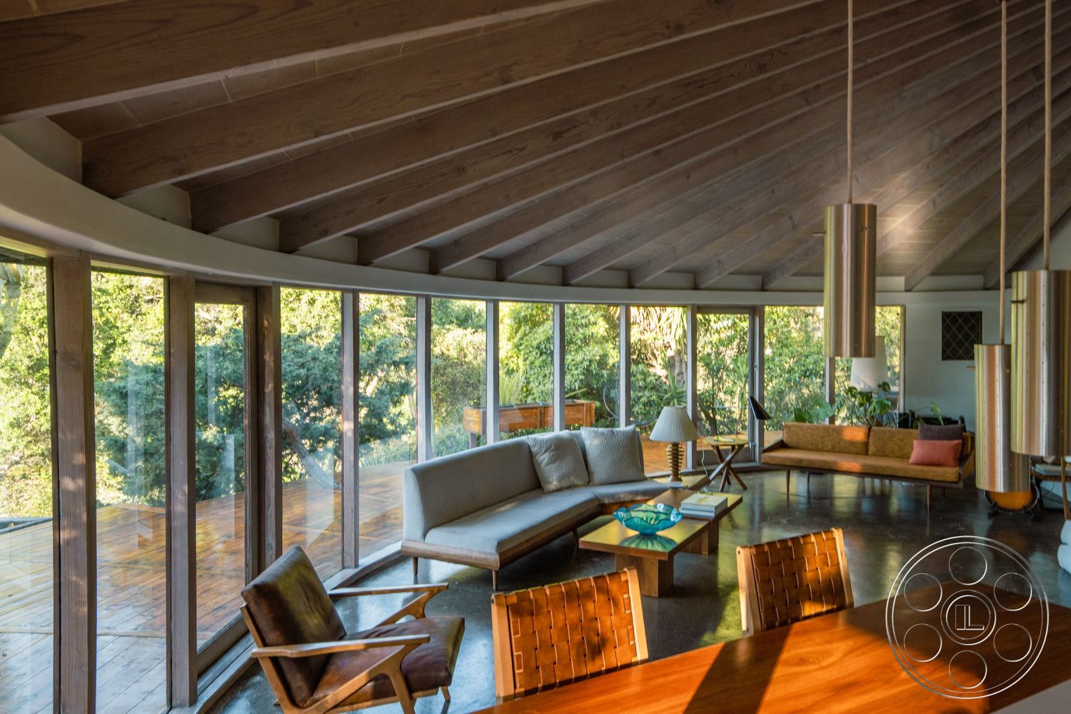 Pad 90210 - mid-century modern architectural style, wood beam ceiling interior, polished concrete floor interior, indoor-outdoor flow interior, minimalist decor interior, wood accent furniture interior, glass wall windows interior, curved living room layout interior, functional furniture arrangement interior, natural light features interior, open concept design interior, deck access from living area interior, exposed wood structural elements interior, urban forest setting view exterior, greenery surrounding view exterior