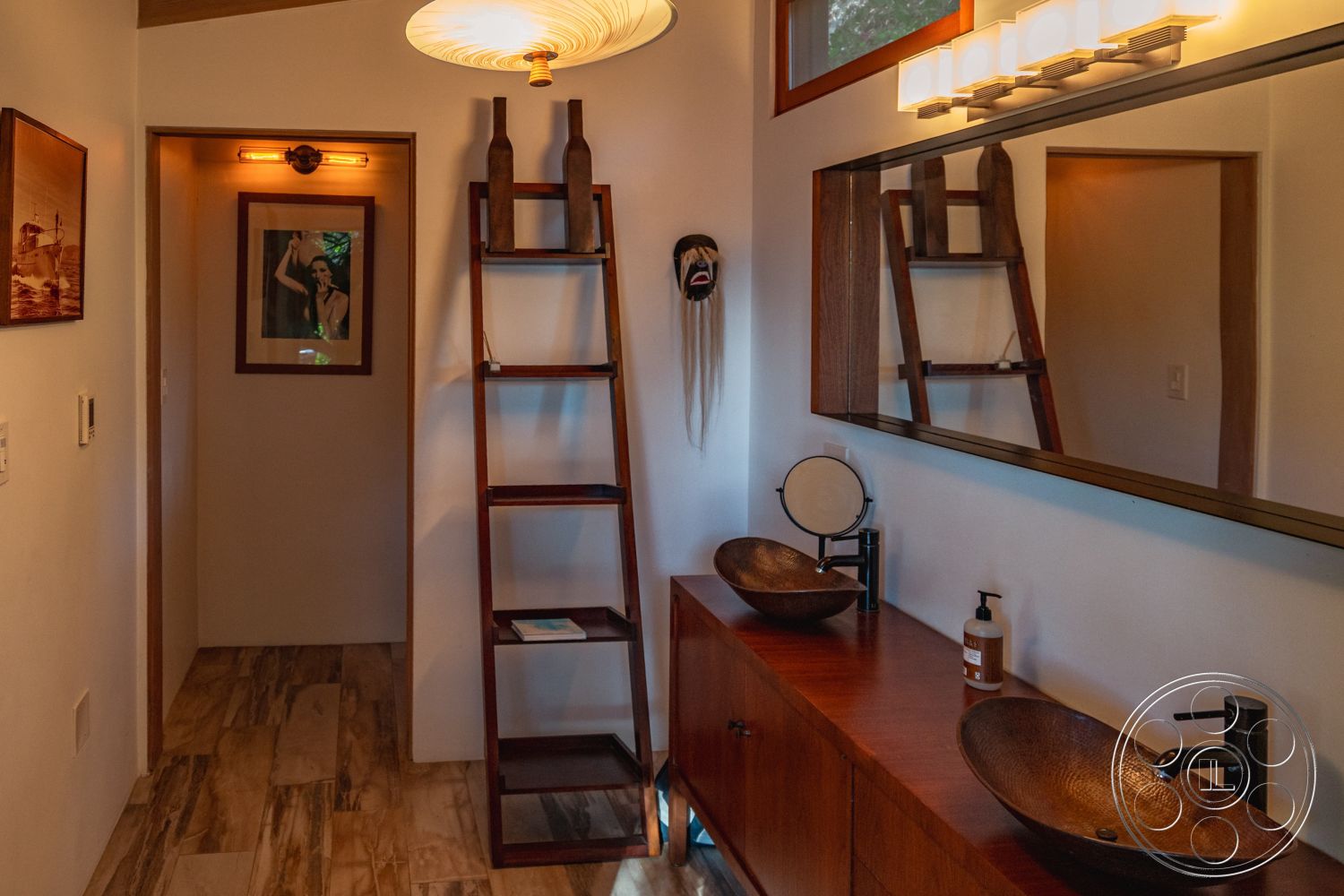 Pad 90210 - warm ambient lighting, natural stone flooring, interior bathroom design, soft natural light from windows, framed artwork on walls, modern wall sconce lighting, minimalist bathroom accessories, large wall mirror, wooden ladder shelf, rustic hardwood vanity, textured ceramic sink bowls, earth-toned wall paint, decorative ethnic mask wall hanging, clean lines and modern aesthetics, open layout with visual depth, wood accents with contemporary style