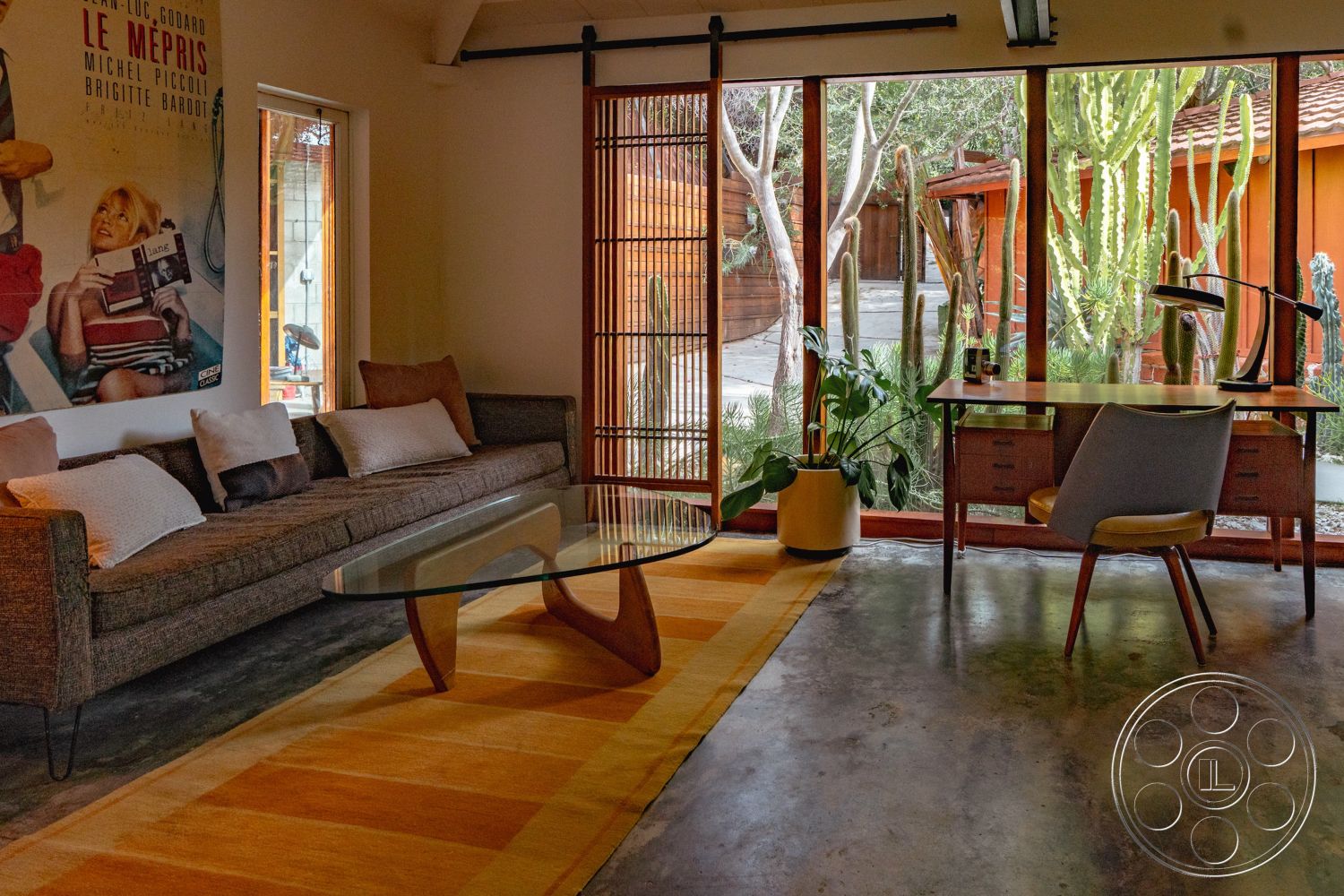 Pad 90210 - textured area rug, open floor plan, glass coffee table, concrete flooring, indoor-outdoor flow, large glass windows, decorative throw pillows, soft color palette, exposed ceiling beams, indoor plant decor, wooden sliding doors, contemporary interior design, natural light abundance, functional workspace area, urban studio setting, mid-century modern sofa, framed artwork wall, retro-style desk, minimalist desk lamp, outdoor cactus garden view