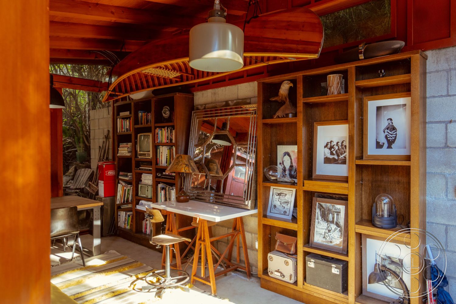 Pad 90210 - polished concrete floor, vintage decor elements, decorative mirrors, cozy reading corner, textured plaster wall treatments, exposed concrete block walls, interior wooden ceiling beams, open shelving for books, hardwood shelving units, vintage office space, metal lamp fixtures, home office workspace, multicolored art wall, rustic wooden desk, black wooden chair, natural foliage view, indoor plants setting, mid-century modern design style, accent wall with framed photos, unique canoe ceiling feature