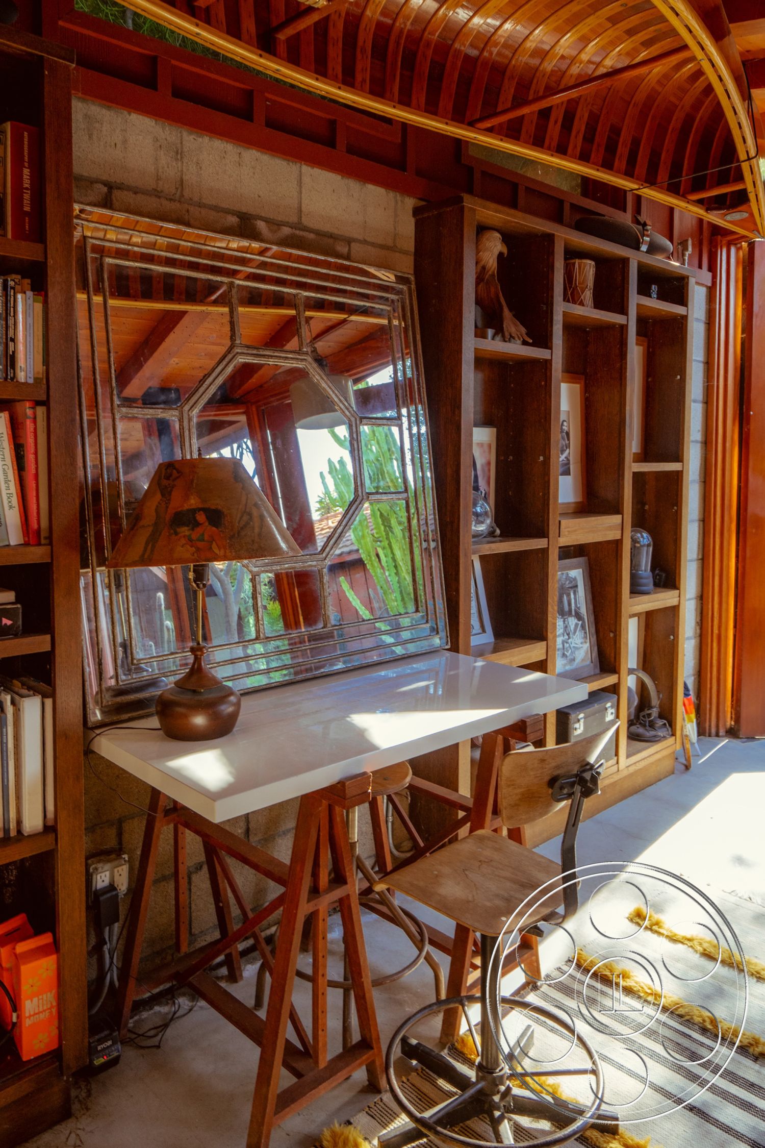 Pad 90210 - indoor plant view, minimalist workspace, exposed concrete block walls, brightly lit environment, rustic wooden accents, natural wood ceiling with curved beams, polished white quartz desk surface, vintage metal bar stool, open bookshelf with wooden shelves, octagonal decorative wall mirror, rugged textile area rug, contemporary desk lamp, large glass window for natural light, studio workspace area, creative office interior, mountain view from workspace, suburban setting with outdoor greenery