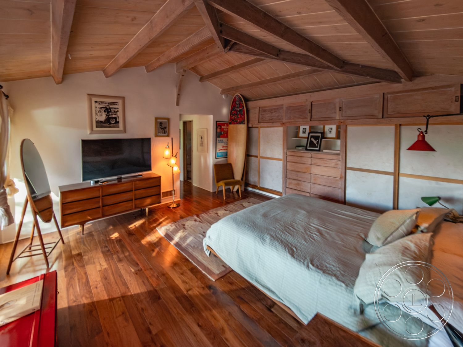 Pad 90210 - textured bedding, decorative wall art, natural light from windows, soft lighting fixtures, earth-toned color palette, flat screen television, contemporary armchair, exposed wood ceiling beams, interior bedroom design, hardwood bedroom floors, open room layout, wood panel accent wall, minimalist bedroom interior, mid-century modern dresser, rustic surfboard decor, simple bedside lamps