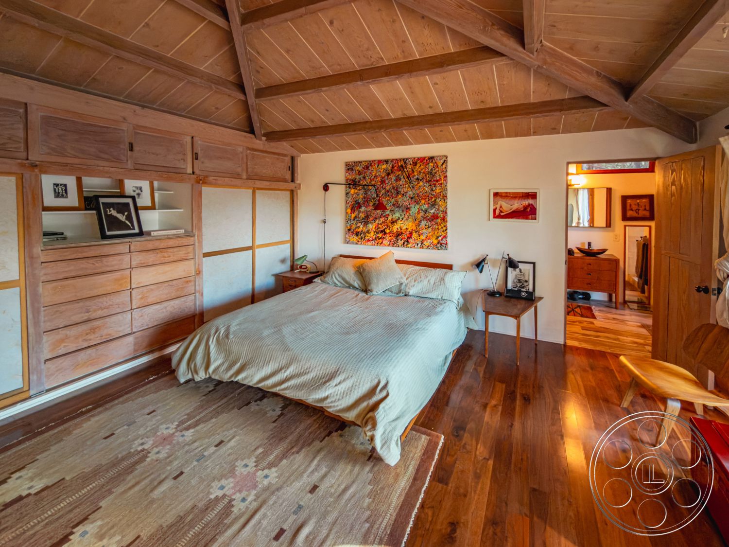 Pad 90210 - decorative wall art, natural light from windows, natural wood paneling, interior bedroom with hardwood floors, minimalist decor style, woven area rug with geometric patterns, exposed wood beam ceiling, framed art on wall, king-sized bed with textured bedding, wooden storage drawers, matching nightstands with lamps, light color palette with warm accents, open door to bathroom area