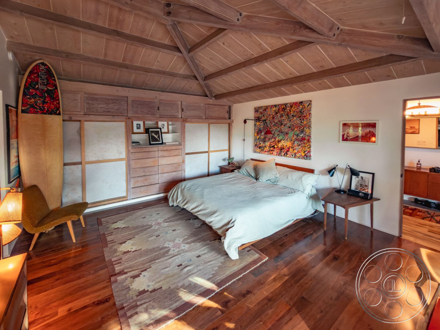 Pad 90210 - open space layout, exposed wood ceiling beams, interior hardwood bedroom floors, soft natural light from windows, textured area rug with geometric patterns, colorful abstract wall art, natural wood paneling accent wall, mid-century modern armchair, wooden dresser with modern design, warm neutral color palette, minimalist bedroom design, contemporary bedside lamps, king-sized bed with linen duvet, sliding wood panel closet doors