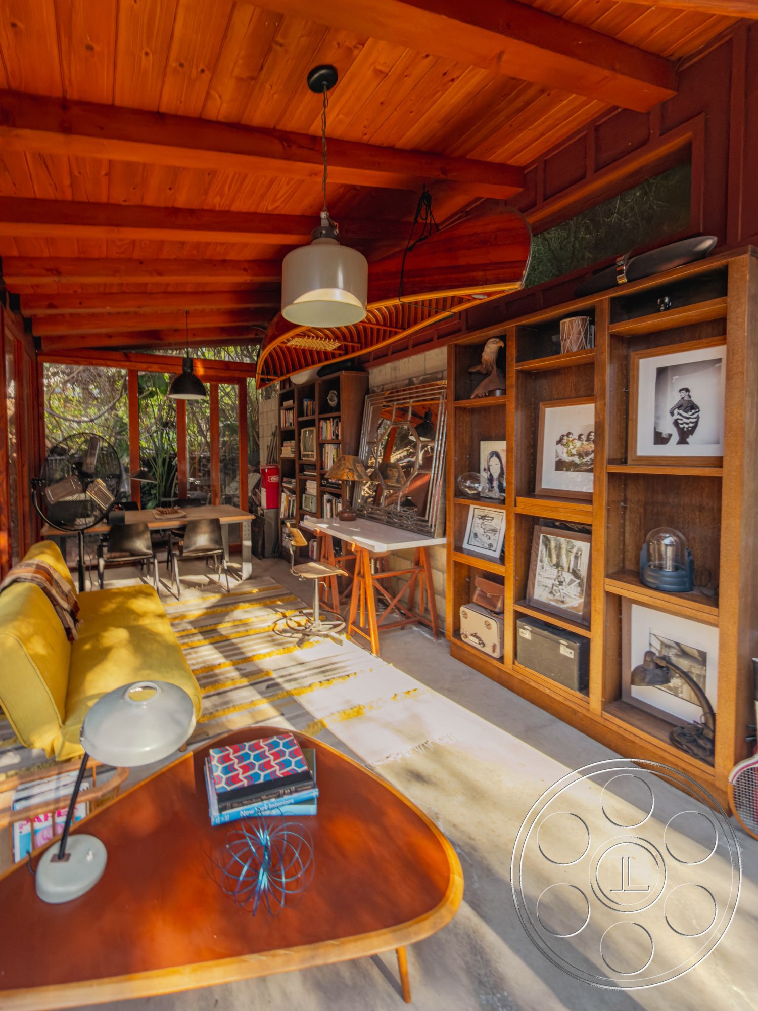 Pad 90210 - textured area rug, open floor plan, concrete floor, black dining chairs, mid-century modern style, photographic wall art, exposed wood ceiling beams, interior living space, modern pendant light fixture, glass window walls, yellow upholstered sofa, light wood paneling walls, wooden coffee table with rounded edges, built-in wooden shelving unit, wooden bar height table, artistic decoration items, outdoor vegetation view