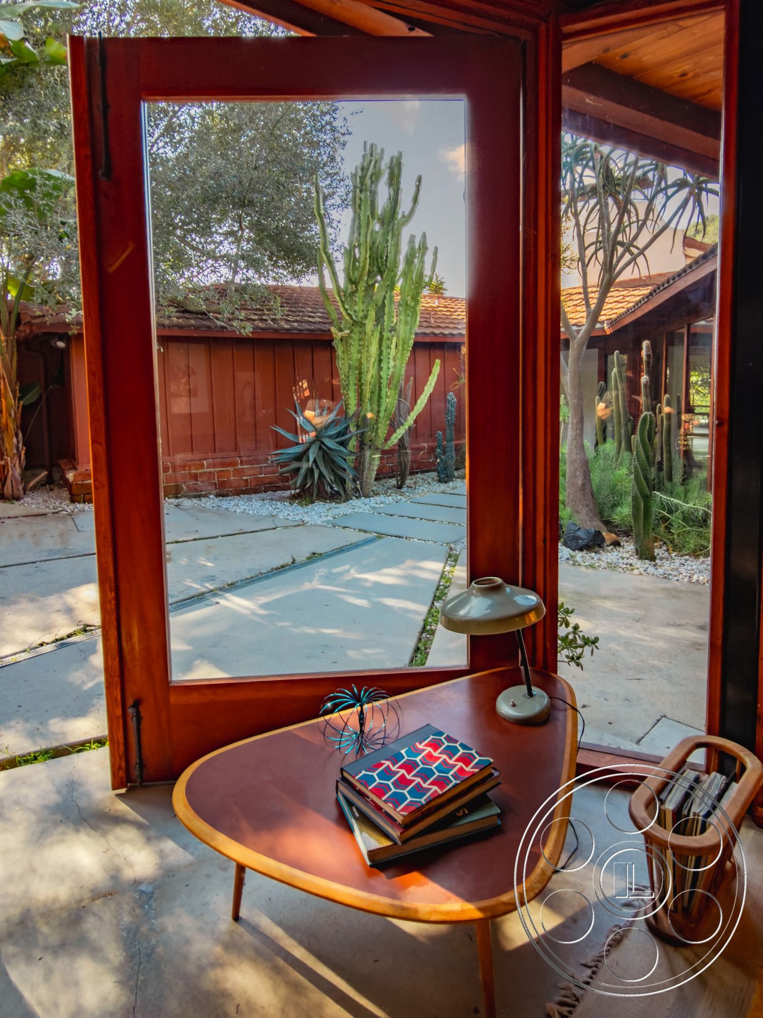 Pad 90210 - exterior, round coffee table, open floor plan, bright natural light, indoor-outdoor flow, sliding glass doors, minimalist furniture, succulent garden, decorative table lamp, courtyard view, wood frame windows, mid-century modern design, modernist architectural style, interior wooden accent walls, textured concrete patio flooring, natural stone landscaping, brown wood window frames, curve-shaped patio layout, desert plant garden, stacked books, urban jungle aesthetic
