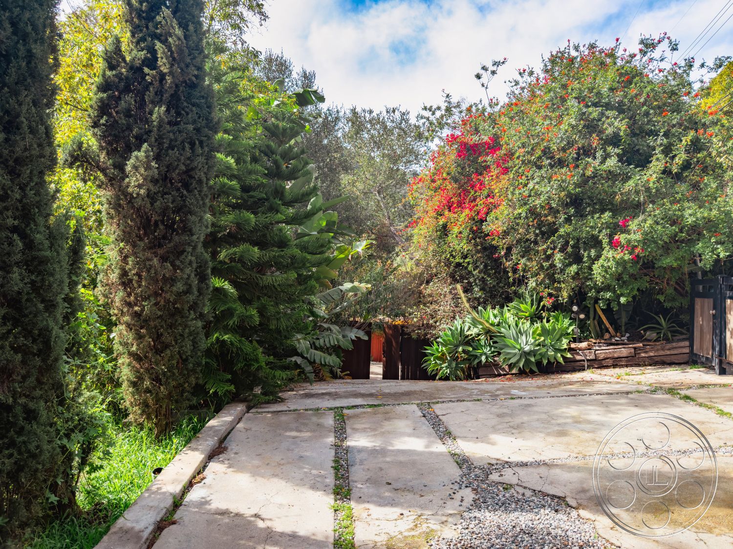 Pad 90210 - exterior driveway made of concrete with gravel inlay, landscaped garden with native shrubs and flowering plants, tall cypress trees providing vertical greenery, lush tropical foliage surrounding the driveway, rustic wooden fence along the property line, natural stone retaining wall for garden bed, paved concrete surface leading to entrance, sun-drenched outdoor setting with scattered clouds, residential setting with a mix of greenery and hardscape, garden patio area with various plant species, urban home with a blend of nature and architecture