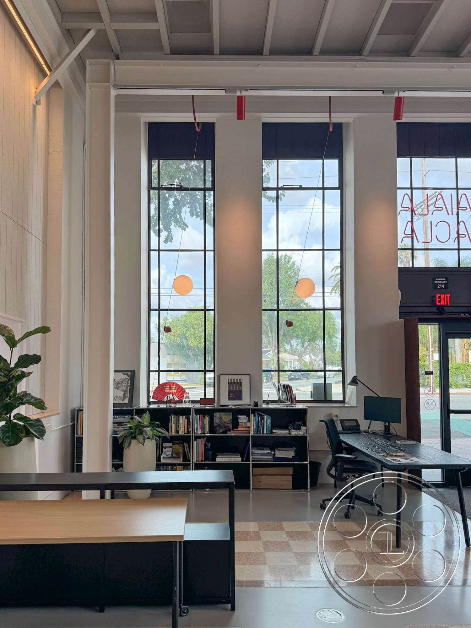 Studio 101 - interior space, functional workspace, contemporary design elements, view of outdoor greenery, black desk lamp, natural light flooding in, potted indoor plants, modern office layout, minimalist workspace, concrete floor tiles, black metal shelving unit, workspace with computer setup, large black-framed windows, open and airy atmosphere, high ceiling with exposed beams, neutral colored walls, checkerboard patterned floor, wooden desk with straight lines, lighting fixtures with red accents