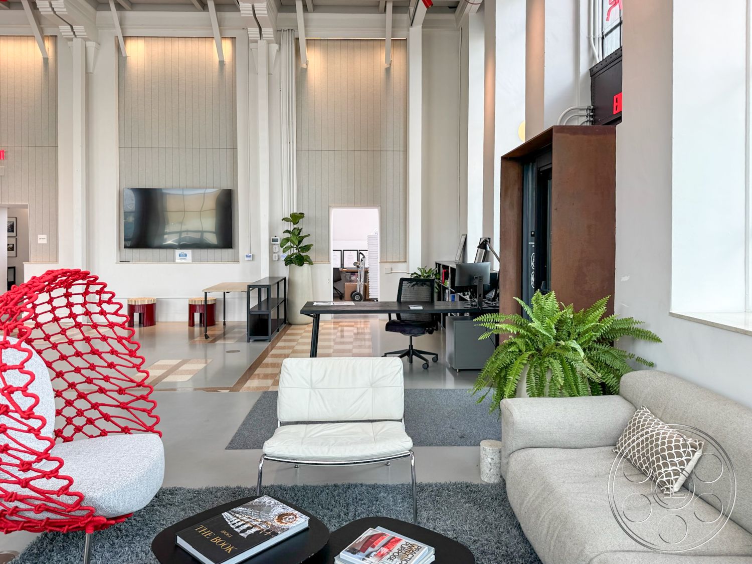 Studio 101 - neutral color palette, minimalist design, concrete floor, textured wall panels, stylish accent pillows, photographic wall art, light gray area rug, glass partition walls, exposed wooden ceiling beams, interior open office space, polished concrete finish, bright natural light from large windows, modern task chairs, modern lounge furniture, fabric upholstered sofa, curved red wire chair, low coffee table with magazines, potted green plant decor, workspace with desk and computer, industrial style design elements