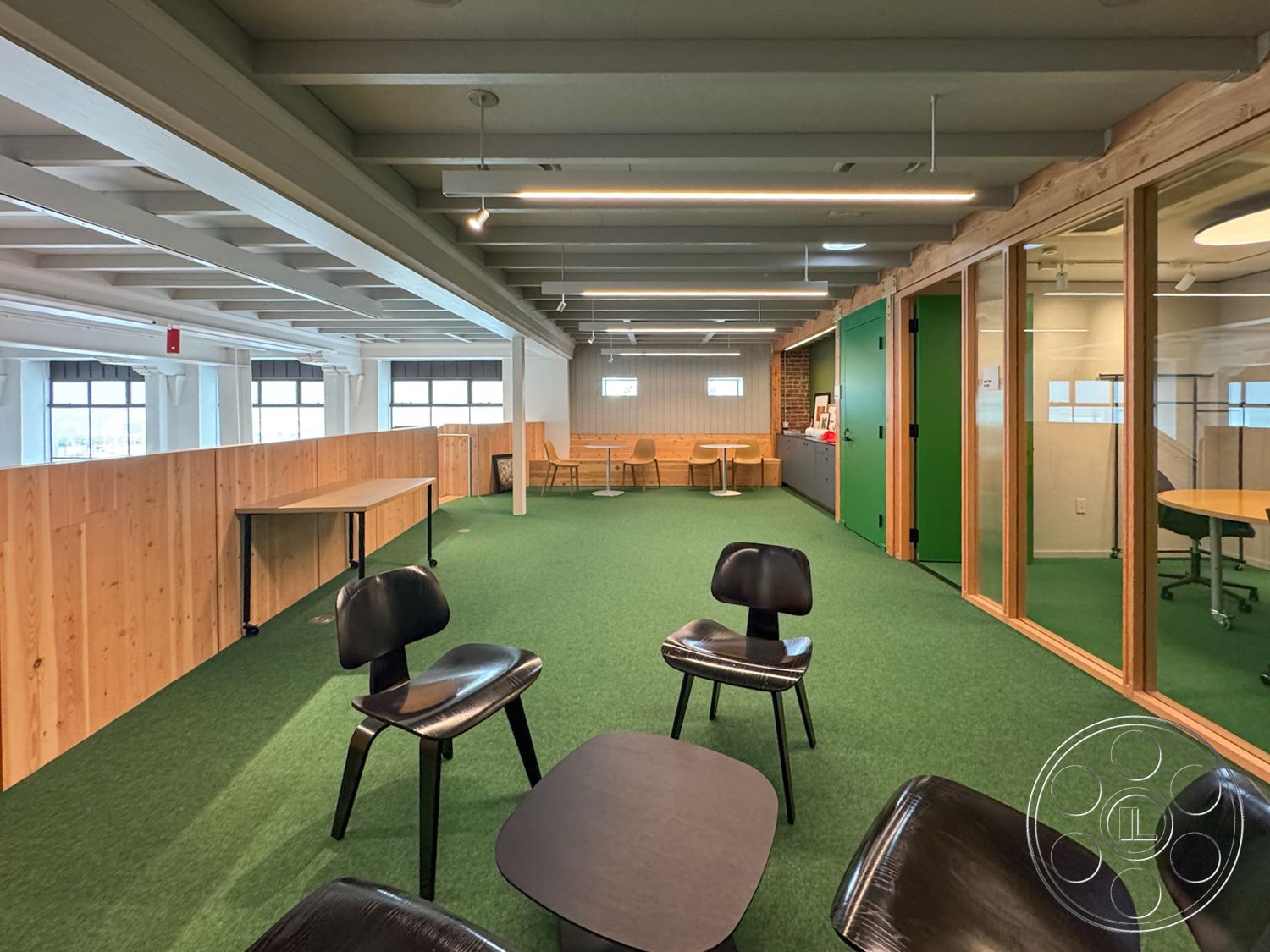 Studio 101 - textured wall accents, floor-to-ceiling windows, open-plan layout, black plastic chairs, exposed wood ceiling beams, glass partition walls, modern minimalist design, natural light exposure, functional office furniture, commercial office environment, interior workspace, light-colored wooden paneling, green indoor carpet flooring, rectangular workspace, contemporary office setting, circular meeting table, wooden workstations, bright green accent walls