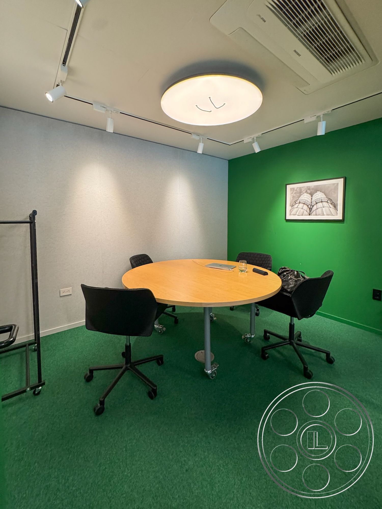 Studio 101 - round wooden table, modern minimalist design, recessed LED ceiling lights, interior conference room, soundproof wall panels, green carpet flooring, circular ceiling light fixture, black office chairs on casters, white plaster ceiling, framed artwork on the wall