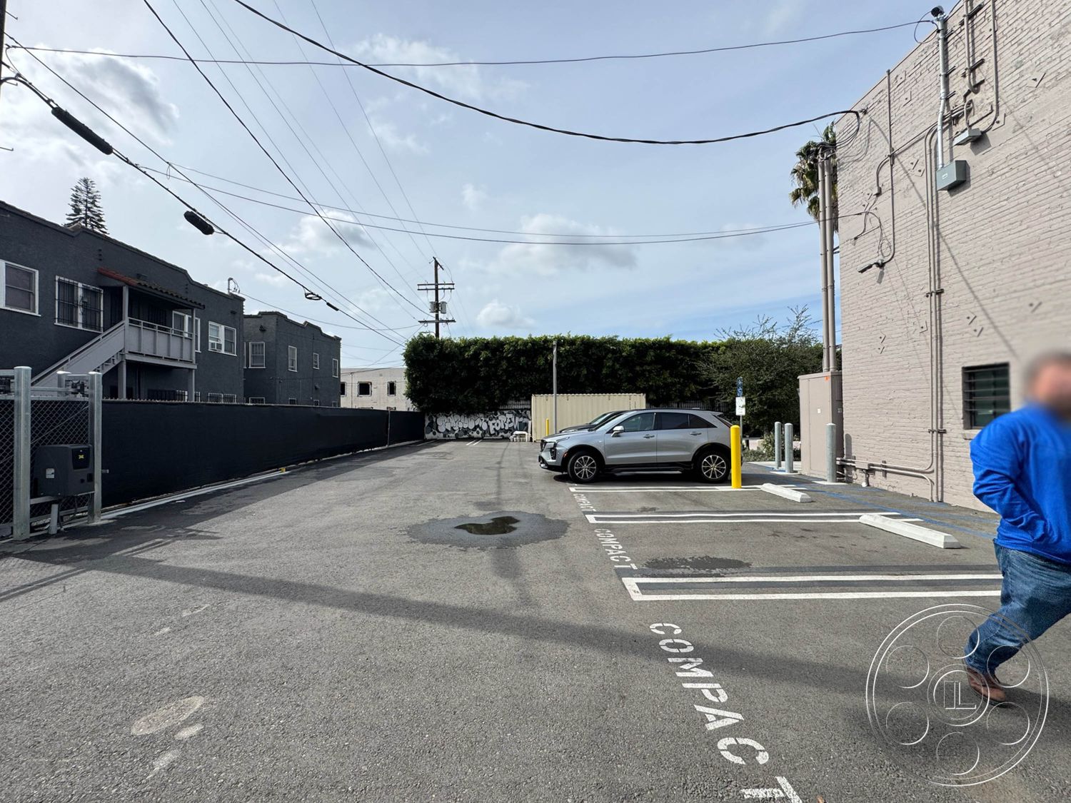 Studio 101 - concrete pavement, urban alleyway setting, exposed brick exterior wall, striped parking lot lines, metal fence enclosure, asphalt driveway surface, utility poles with wires, residential backlot, landscaped hedge boundary, modern SUV parking space, gray apartment building, cloudy daytime sky, compact car parking area, concrete footpath, interior alleyway view