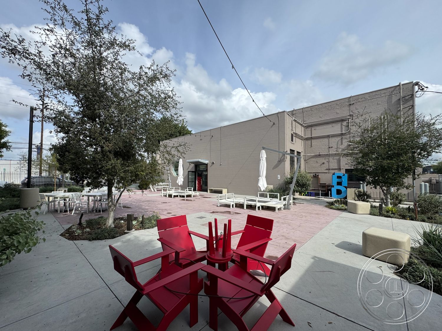 Studio 101 - exterior, open space design, surrounding greenery, casual dining area, functional outdoor space, shade umbrellas, concrete patio flooring, landscaped garden beds, urban outdoor setting, pink patterned paver stones, red wooden outdoor chairs, round table with red accents, modern site furnishings, textured planter boxes, large commercial building facade, gray painted brick walls, lantern-style string lights, urban outdoor lounge area