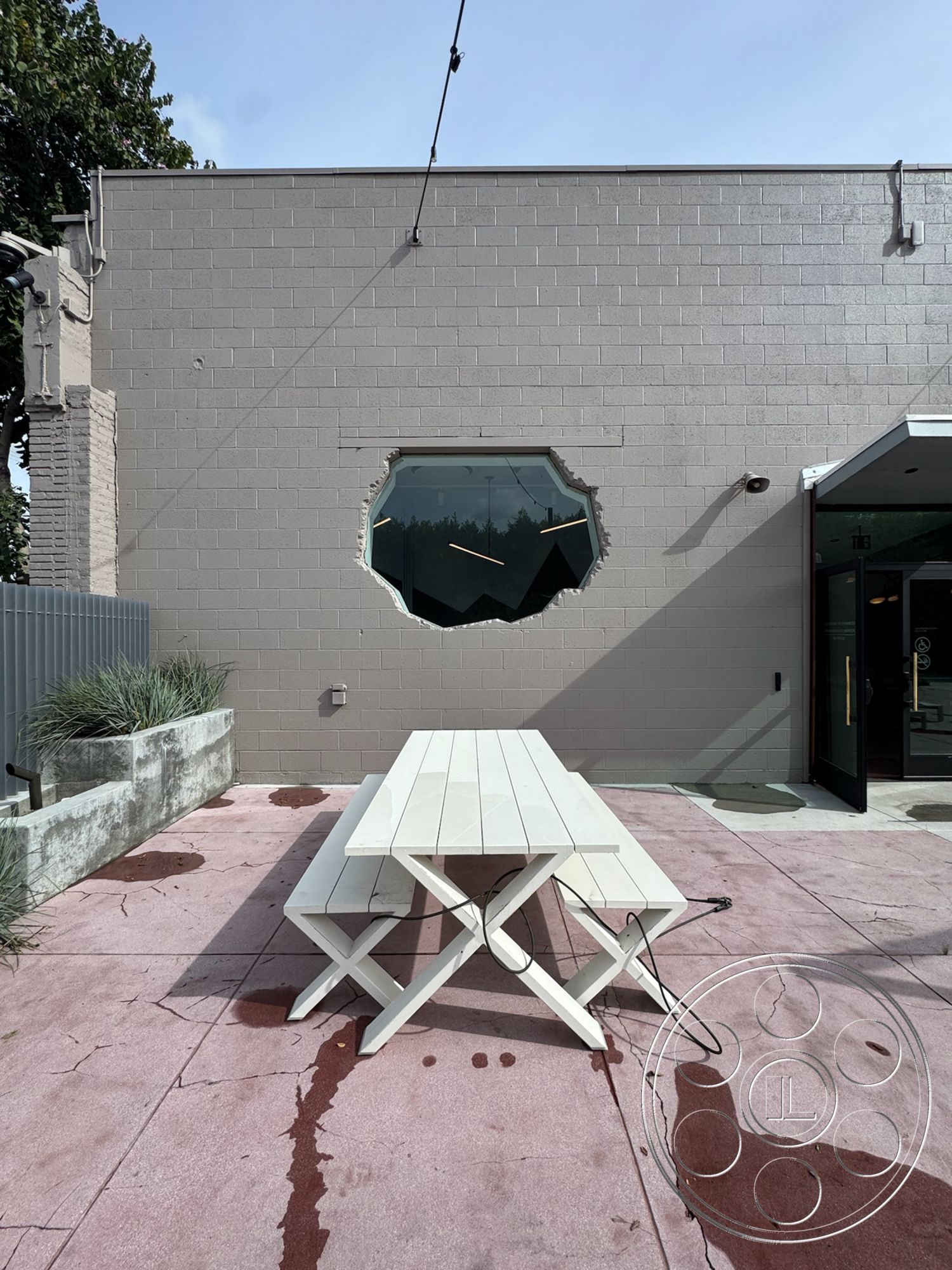 Studio 101 - exterior wall treatment - painted concrete block wall, octagonal window with modern framing, outdoor picnic table - white wood construction, textured patio flooring - pink stained concrete, landscaped planter with concrete borders, urban outdoor setting with greenery, open air dining area, interior/exterior contrast with modern design elements, industrial style architecture with minimalist features, natural light exposure through large window