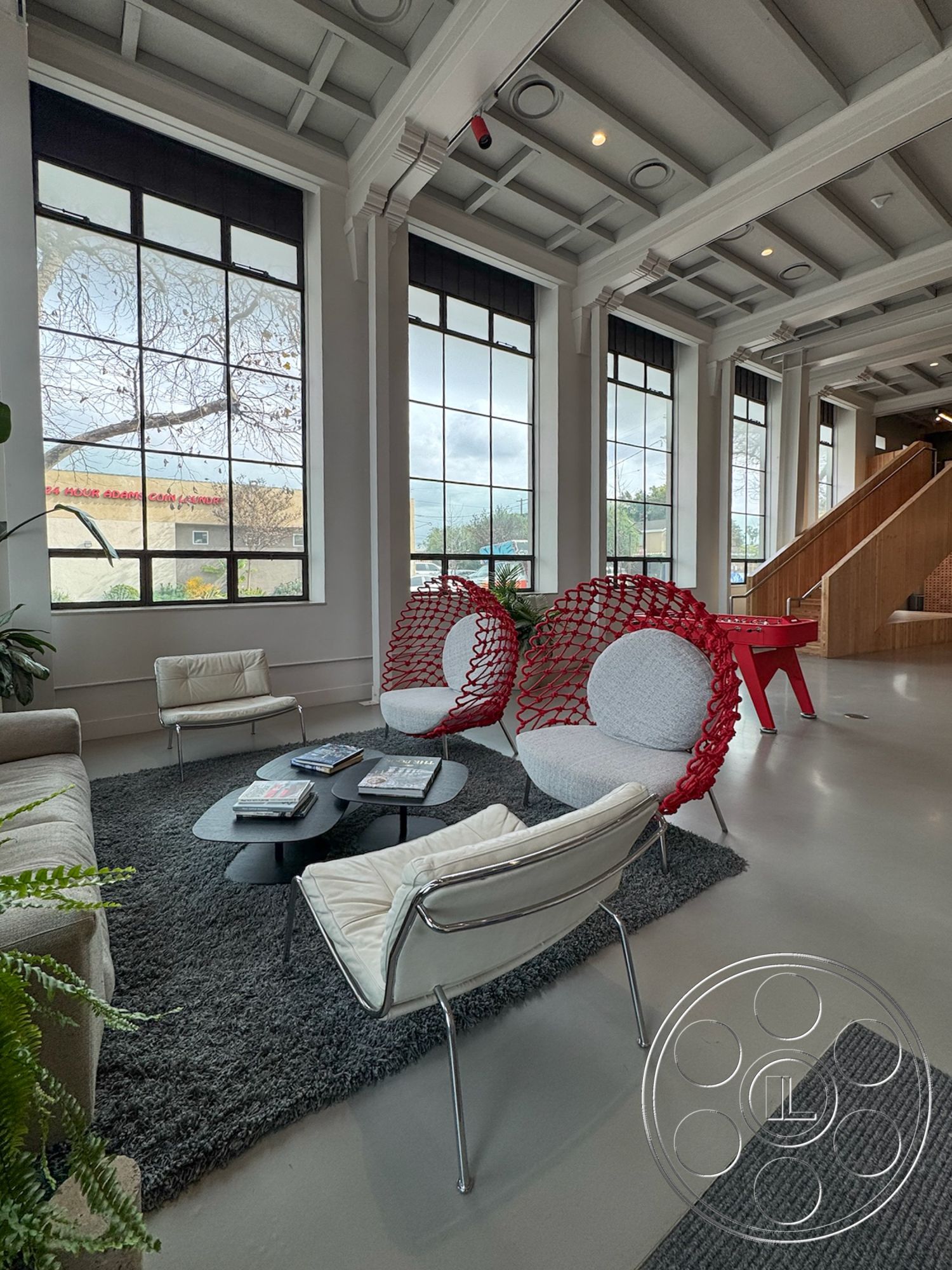 Studio 101 - minimalist decor, textured area rug, large glass windows, floor-to-ceiling windows, green indoor plants, bright natural lighting, exposed wood ceiling beams, open floor plan living area, urban interior setting, modern round coffee table, industrial style staircase, neutral colored walls, light gray polished concrete flooring, red plastic rope chairs, contemporary white upholstered chairs