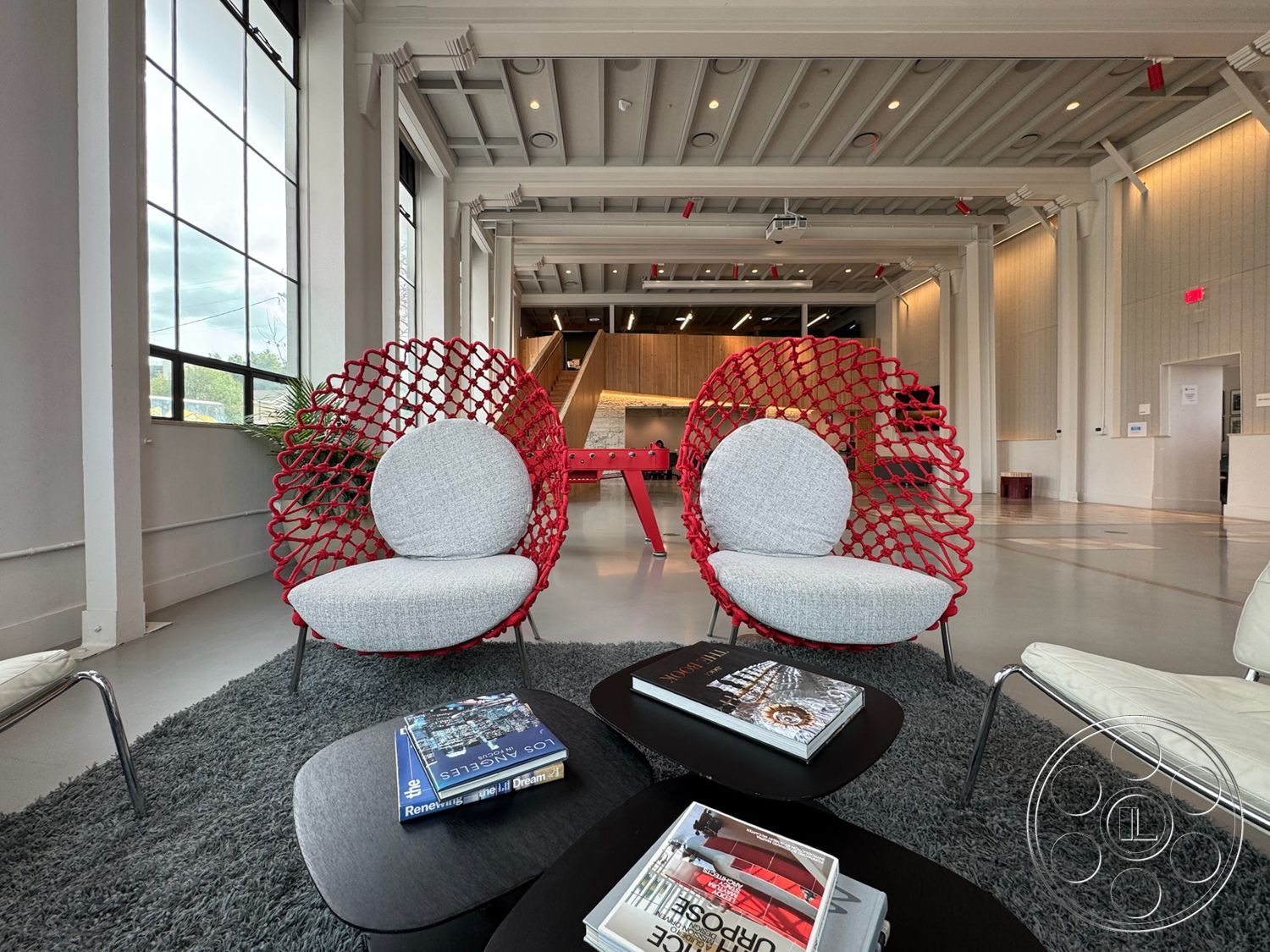 Studio 101 - minimalist aesthetic, large industrial windows, exposed wood ceiling beams, open floor plan layout, white painted walls, soft area rug flooring, architectural lighting fixtures, urban loft setting, creative workspace environment, contemporary decor elements, concrete floor with polished finish, red woven lounge chairs, interior modern industrial design, gray upholstered seating, black coffee table with rounded edges, green plants as interior accents, multifunctional furniture