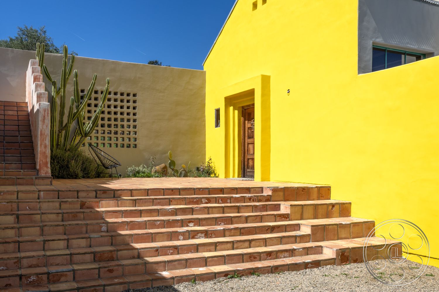 Casa del Rey - rectangular planters, gravel ground cover, exterior stucco walls, textured plaster wall, green foliage accents, desert landscaping with cacti, yellow painted stucco, terracotta tile steps, wood front door with decorative details, stepped entryway design, Bohemian
