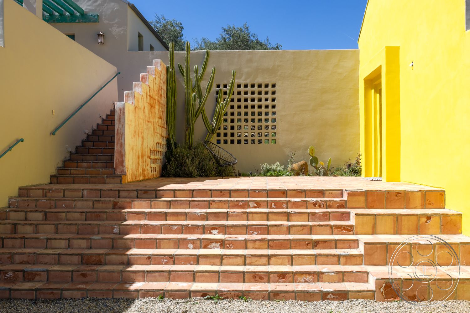 Casa del Rey - natural stone accents, desert landscape, stucco walls, minimalist outdoor space, urban garden patio, built-in seating area, sunlit outdoor area, courtyard setting, geometric stair design, terracotta stair treads, green metal handrails, textured concrete patio floor, exposed concrete block wall, colorful yellow exterior wall, cactus landscaping