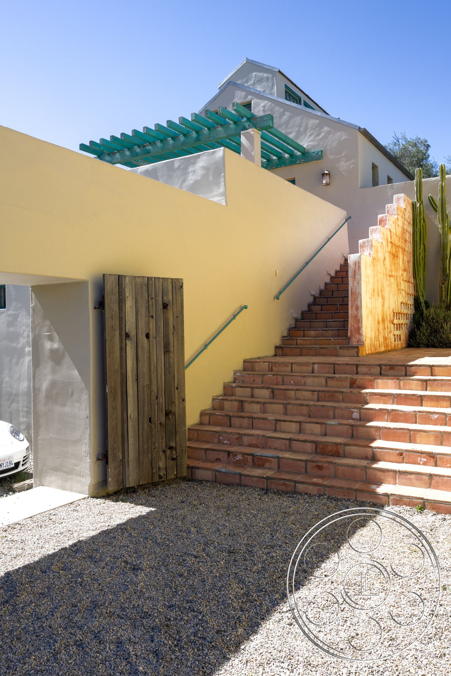Casa del Rey - exterior, low-maintenance landscaping, sloped terrain, gravel pathway, modern architectural style, desert landscaping, wooden gate, stucco wall finish, brick staircase, pergola with wooden beams, rustic wooden accents, steel handrail, steps with textured brick, bright sunlit entrance