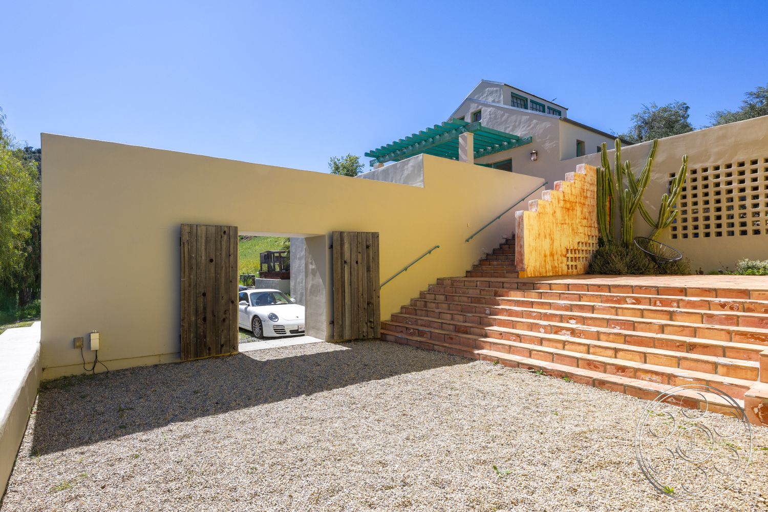 Casa del Rey - modern architectural style, textured plaster walls, gravel driveway, exterior concrete walls, surrounding hillside view, exterior wooden gate, decorative concrete steps, stucco wall treatments, light-colored concrete with a smooth finish, multi-level outdoor staircase, wooden railing on stairs, geometric stepped design, desert planting with cacti, green pergola, urban setting with landscaped elements
