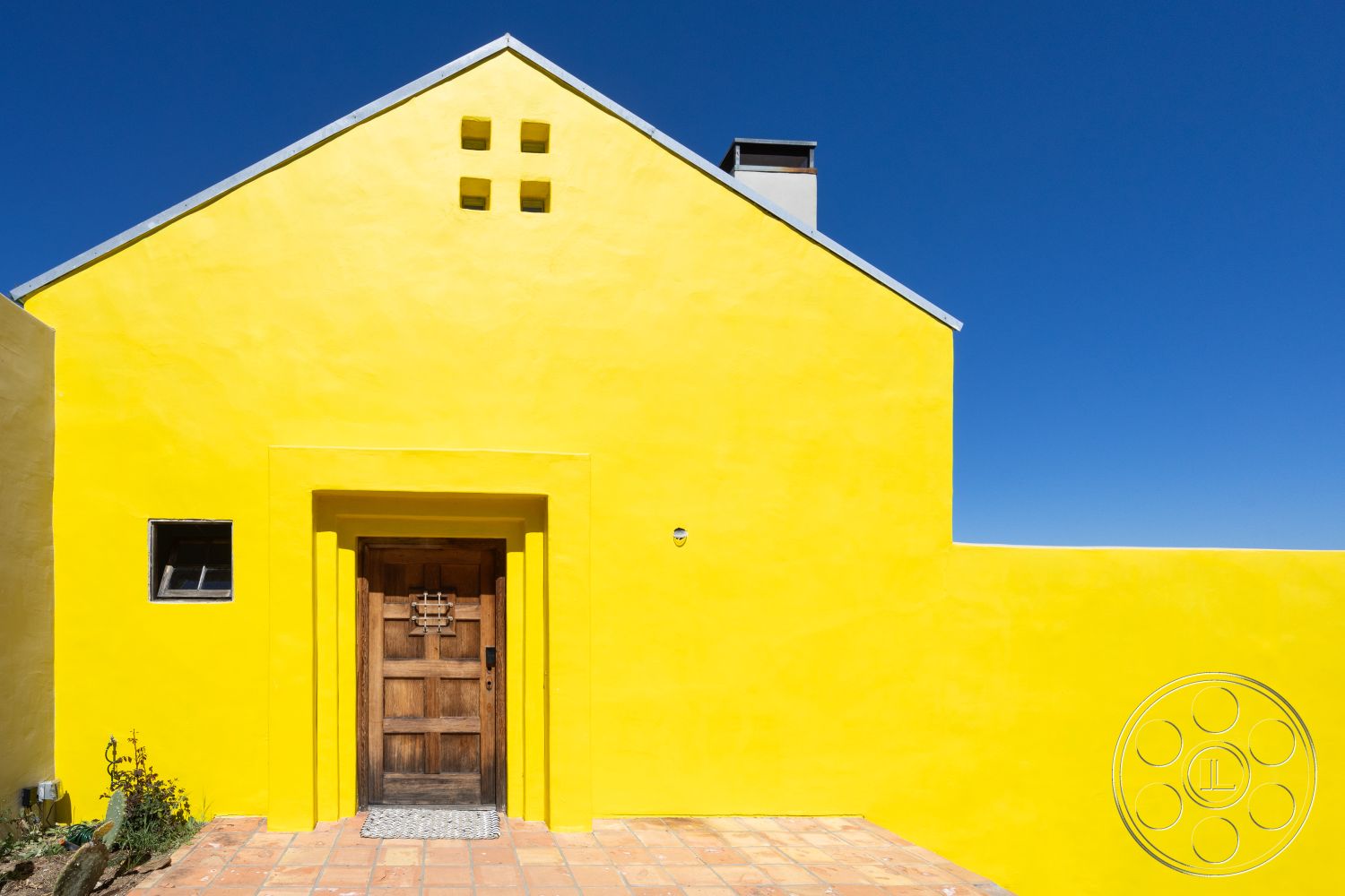 Casa del Rey - exterior, inviting entrance, single-story house, wood front door, desert setting, small square windows, gabled roof shape, minimalist architectural style, symmetrical façade, clear blue sky background, yellow stucco wall, terracotta tile pathway