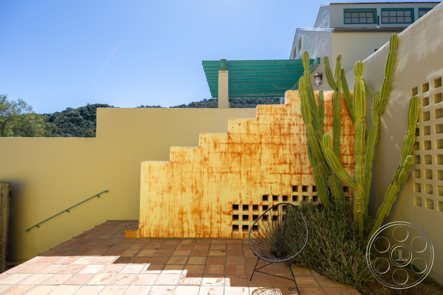 Casa del Rey - exterior patio with terracotta tile flooring, rusted metal stair feature, stucco wall with a warm beige finish, cacti plants in landscaping, black metal chair in outdoor seating area, green wooden pergola atop stairs, textured concrete wall with cut-out patterns, landscape view with surrounding hills, architectural design featuring modern forms, outdoor living space with minimalist aesthetic