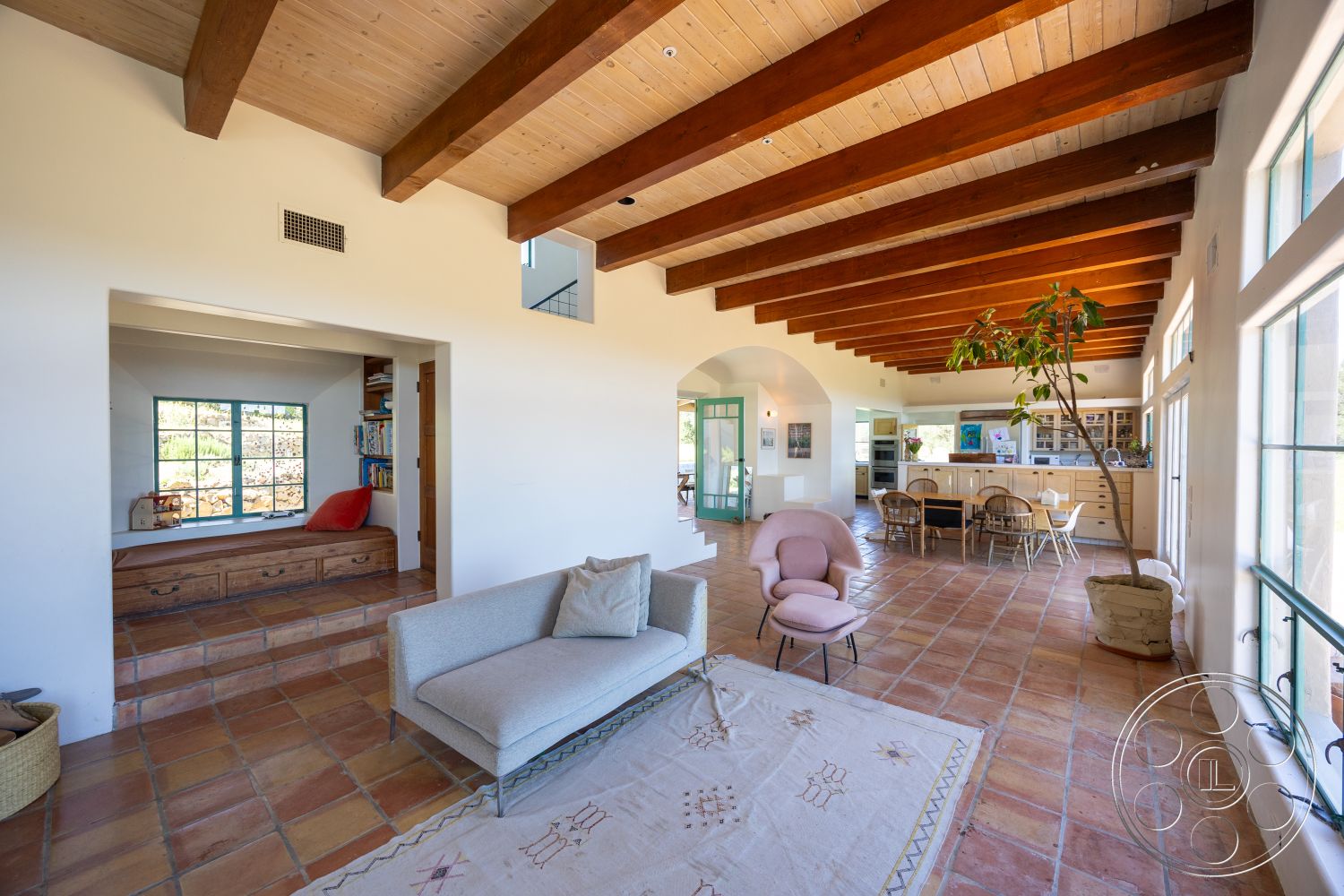 Casa del Rey - neutral color palette, casual dining space, open concept living space, arched doorways, rustic wood coffee table, exposed wood ceiling beams, large rectangular windows, built-in bookshelf nook, suburban home interior, sunlit garden view, minimalist furniture arrangement, spacious communal area, terra cotta tile flooring, natural light-filled interior, stylish lounge chair, comfortable fabric sofa, light plaster walls, ultra-modern kitchen area, friendly living room setting, earth-toned area rug