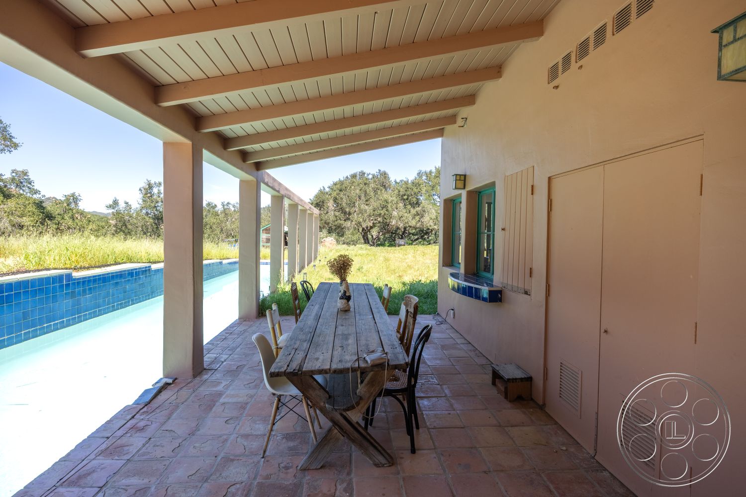 Casa del Rey - natural light, wood dining table, rectangular swimming pool, green landscape view, stucco walls, terracotta tile flooring, outdoor lounging space, overhang roof, exposed wood ceiling beams, exterior patio area, blue tiled pool border, modern outdoor dining, gentle slope garden, rustic-style outdoor design
