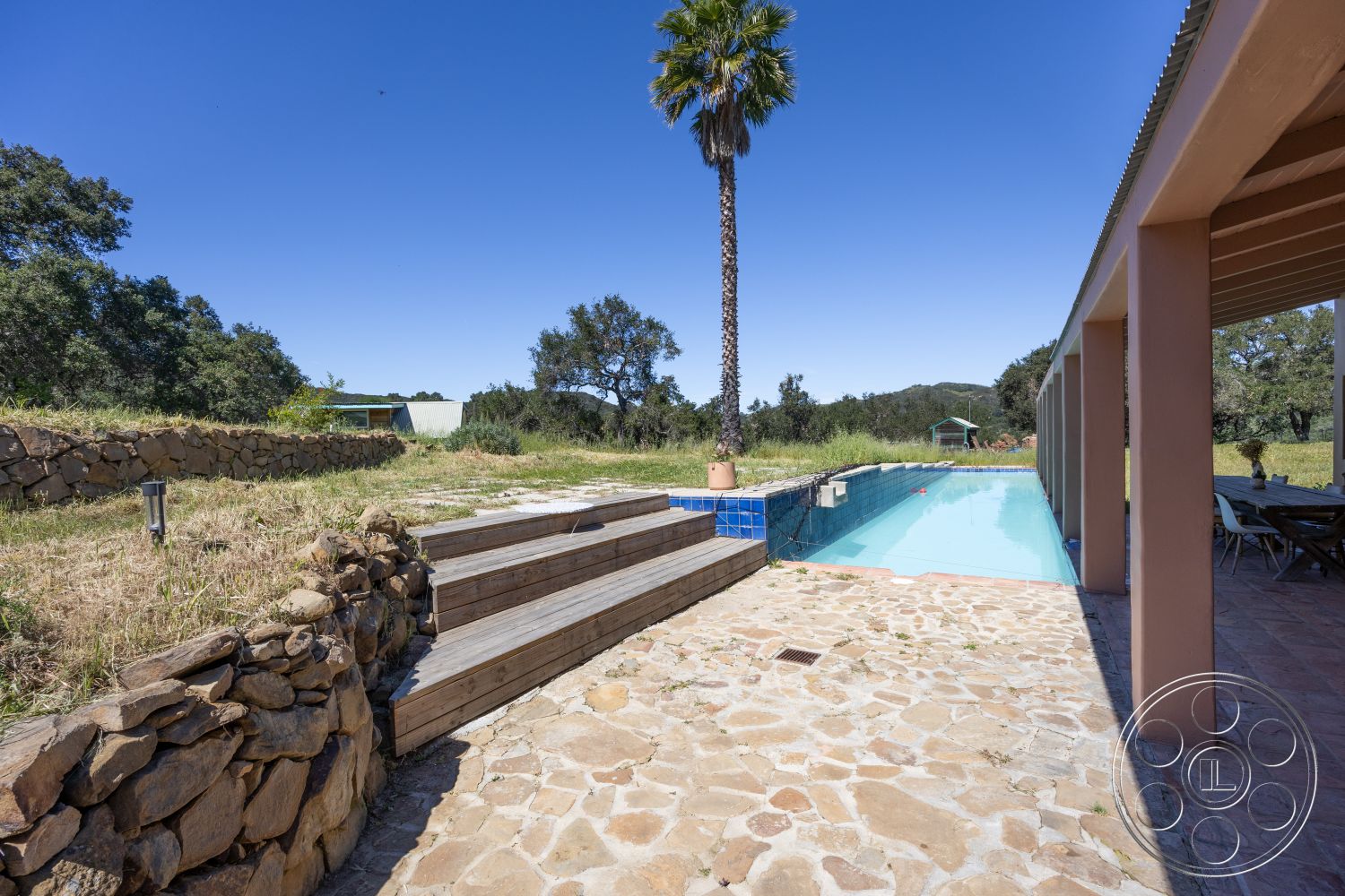 Casa del Rey - textured concrete walls, outdoor dining area, mountain view, rectangular pool, stone pool deck, sunny exterior, palm tree landscaping, natural stone patio flooring, modern outdoor living space, landscaped grassy area, exposed stone retaining wall, wooden pool steps, rural outdoor setting