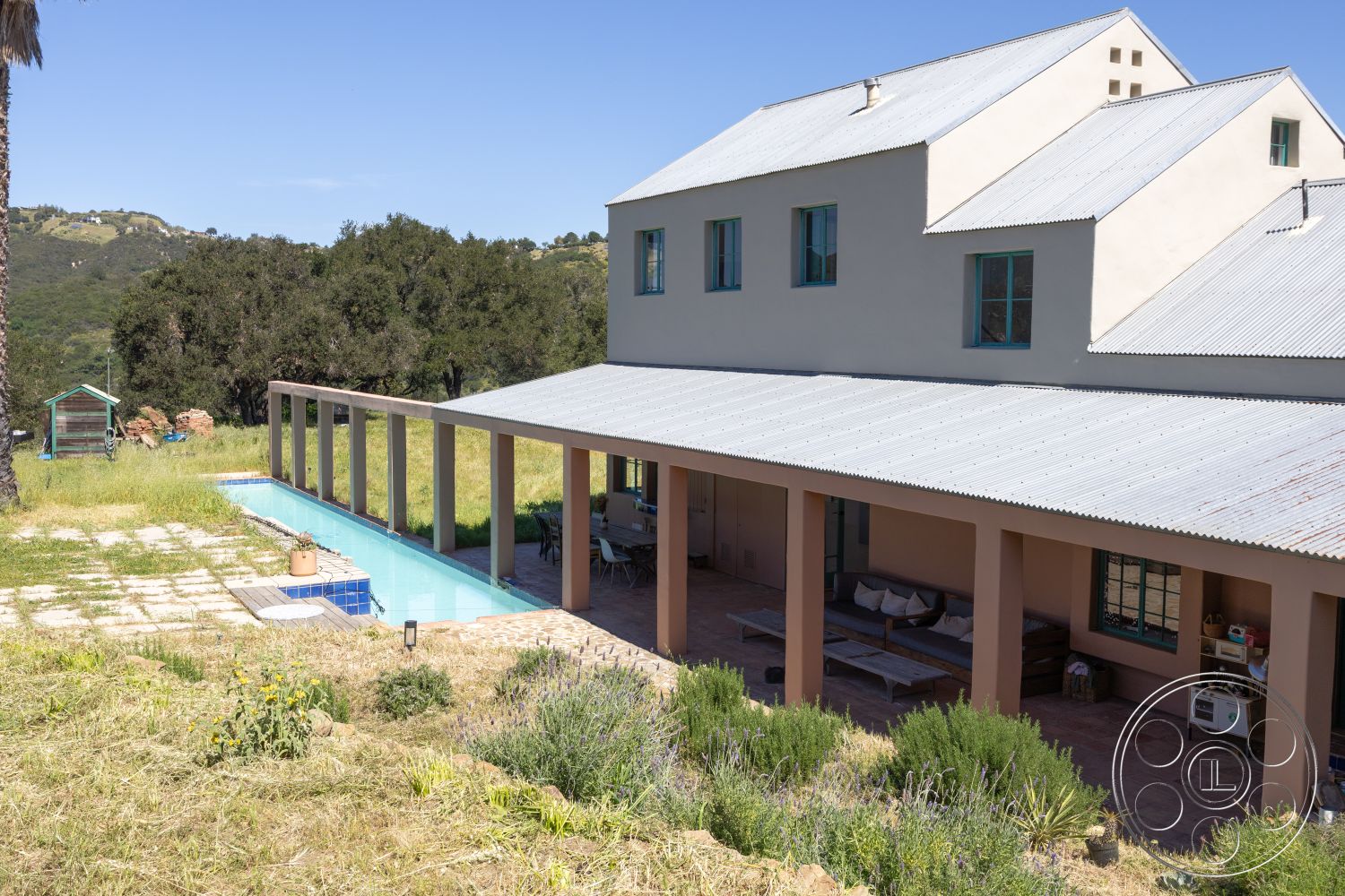 Casa del Rey - rectangle swimming pool, exterior concrete pool border, textured plaster wall, landscaped garden beds, outdoor lounge seating, hardwood patio flooring, interior exposed beam ceiling, surrounding hillside view, modern farmhouse architectural style, metal corrugated roof, steel frame outdoor structure, sloped grassy landscape setting, interior living room lounge area, outdoor dining space under pergola, outdoor rustic tile flooring, interior window with blue trim, concrete patio base, urban-countryside transition environment