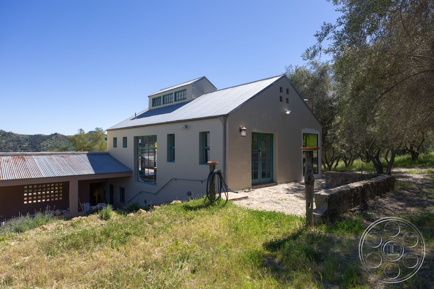 Casa del Rey - exterior, landscape view, metal roof, modern farmhouse style, stucco wall finish, natural landscape setting, stone patio flooring, greenery surrounding, exposed rafters, wooden door with glass panels, built-in outdoor seating, windowed gable end, mudroom entry, sloped terrain location, rustic accent features