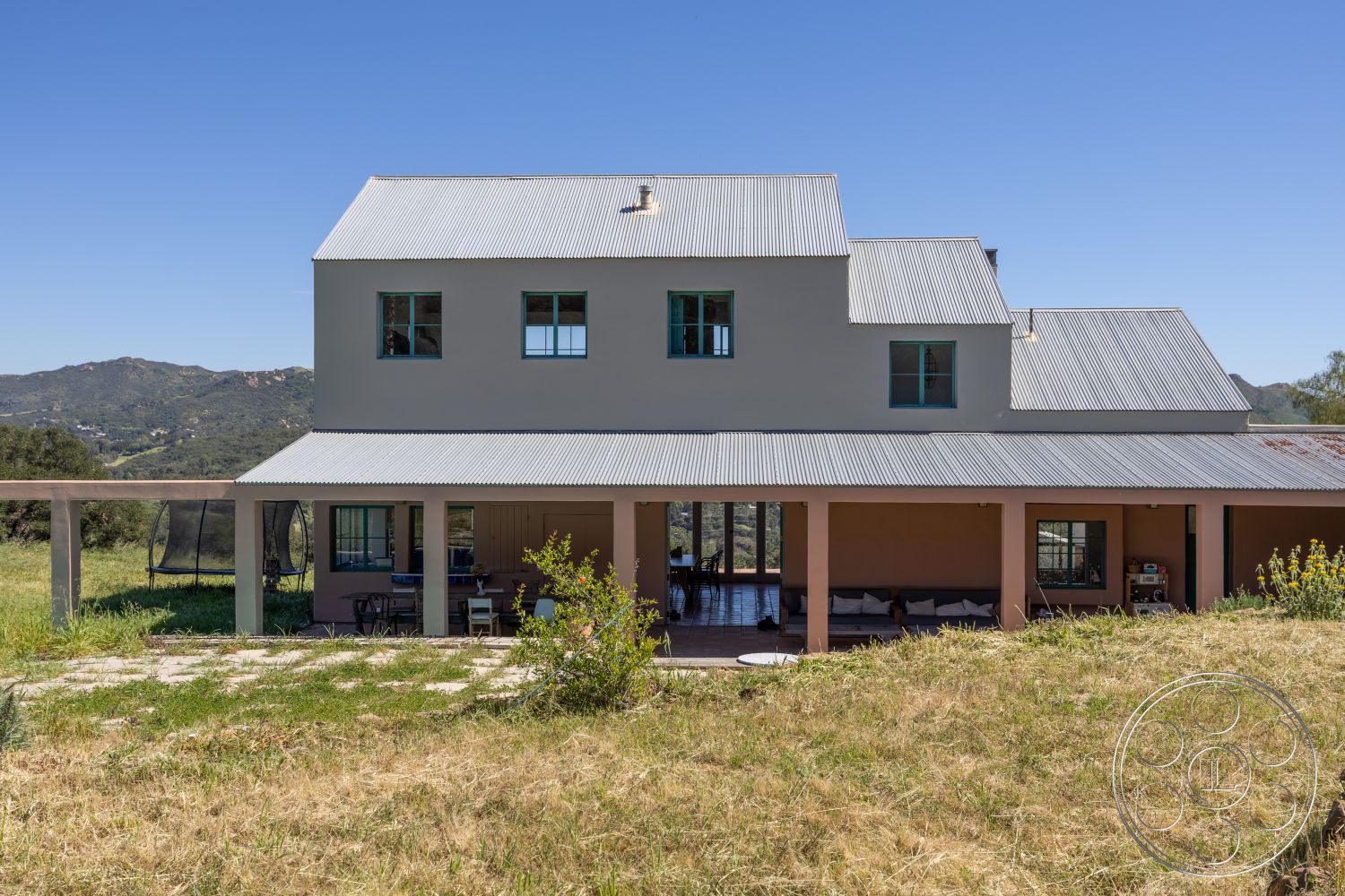 Casa del Rey - exterior, large glass doors, stone pathway, covered porch, mountain view, metal roof, modern farmhouse style, stucco exterior walls, rural hillside setting, open outdoor seating area, glass windows with wooden frames, two-story home design, surrounding grassy landscape, dual-pitched roof, pillars supporting porch