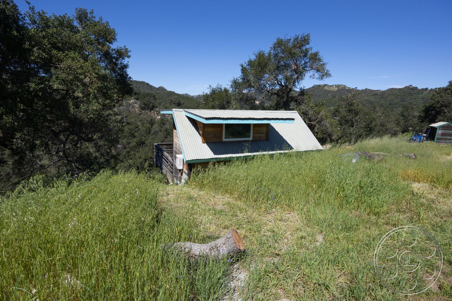 Casa del Rey - landscaped surroundings, natural landscape, mountain view, outdoor rustic charm, interior natural light, sloped roof design, exterior wooden deck, rural hillside setting, minimalist architectural style, metal roof panels, eco-friendly construction, wooden exterior siding, large window with wooden frame, surrounding forest greenery, open grass area, single-story cottage, simple linear forms, asymmetrical roof design