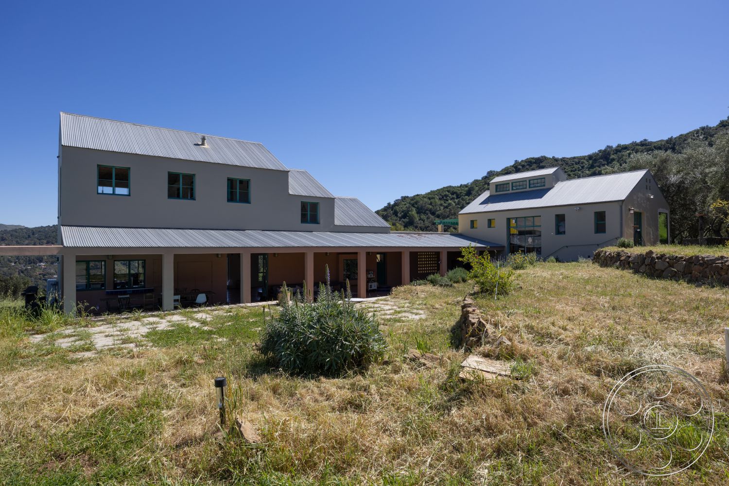 Casa del Rey - large windows for natural light, concrete stepping stones, landscaped garden area, exterior stucco walls, open patio area, minimalist architectural style, exterior metal roof, exterior stone boundary wall, rectangular farmhouse layout, wooden porch supports, exposed beams under the porch, rural hillside property, weekend retreat in natural setting, surrounding greenery and hills, asymmetric roof lines, porch seating area, modern construction techniques, multifunctional outdoor space