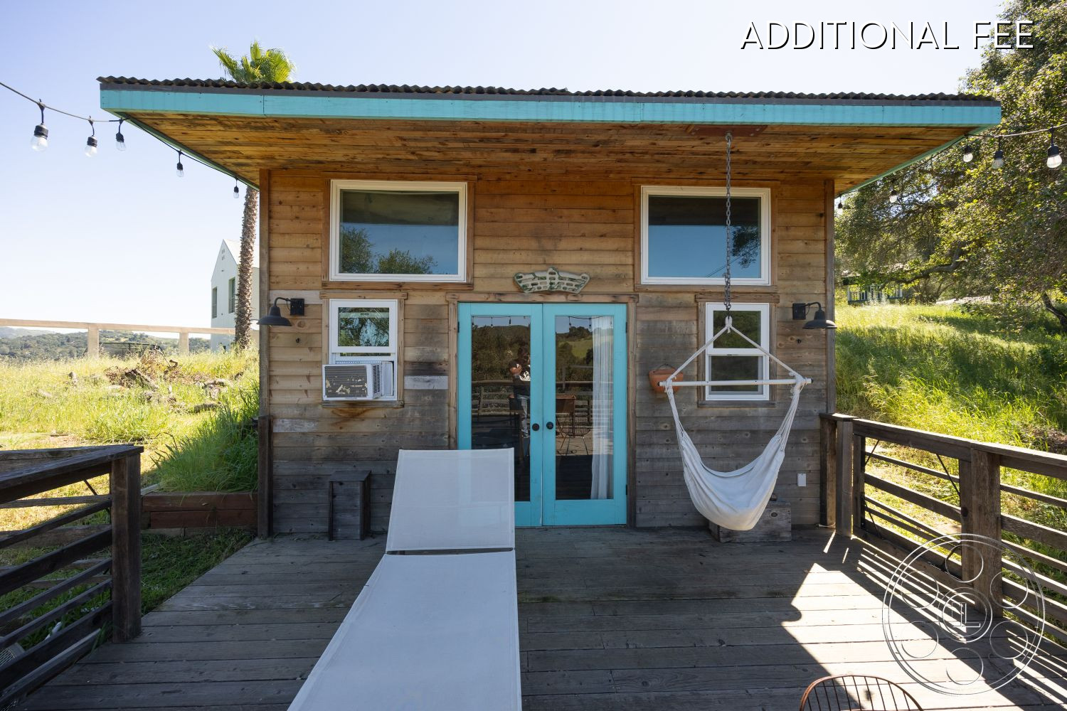 Casa del Rey - wood deck patio flooring, large windows for natural light, exposed wood beam details, minimalist outdoor seating, hammock hanging chair, casual outdoor relaxation area, flat roof with overhang, weathered wood siding exterior, bright turquoise double doors, string lights for outdoor ambiance, open grassy landscape setting, rustic modern architectural style, modern exterior light fixtures, suburban countryside location