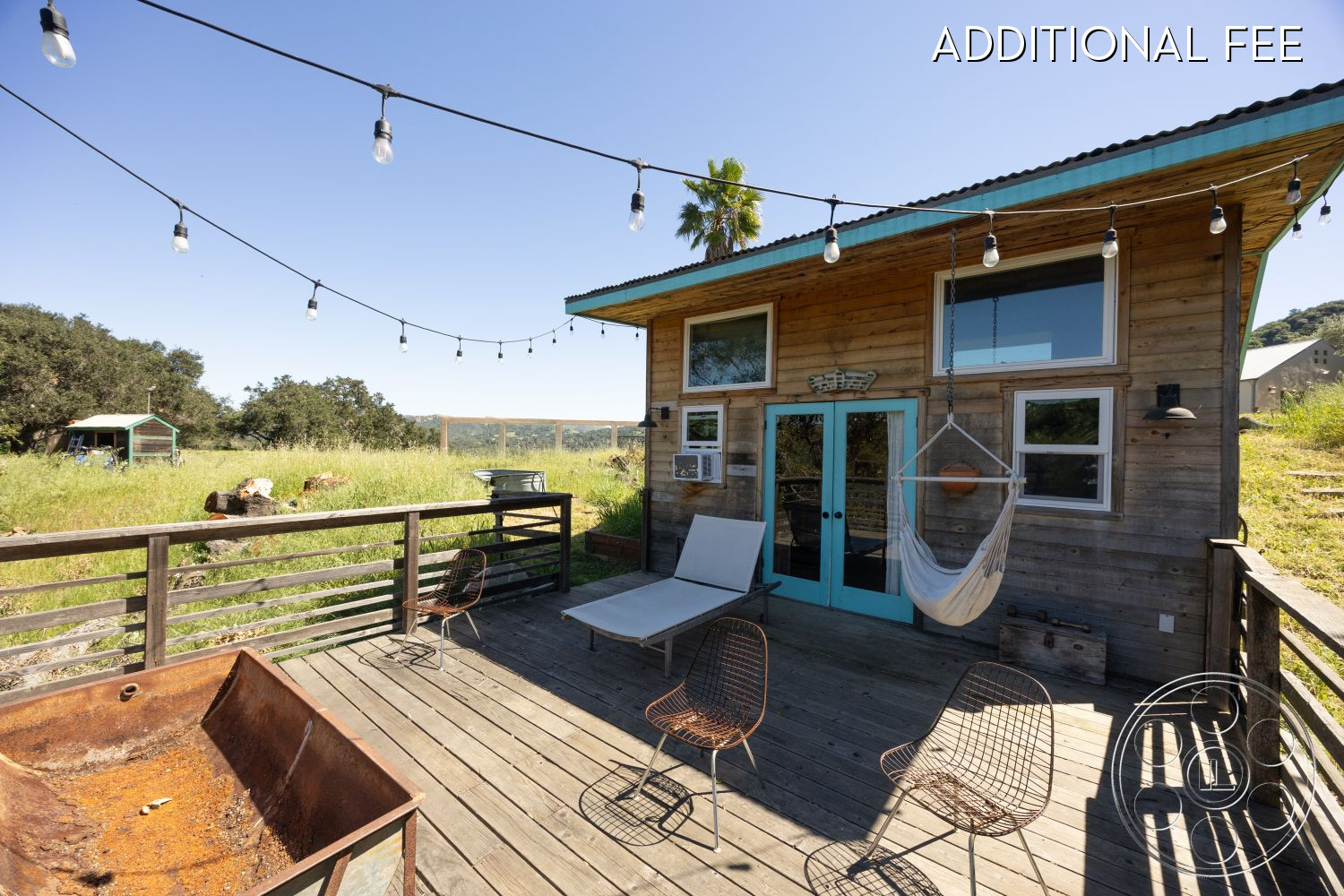 Casa del Rey - modern farmhouse style, wood deck patio, sloped hillside location, exterior wood siding, rustic outdoor fire pit, hammock hanging chair, teal front door, exposed wood deck flooring, metal wire chairs, string lights outdoor, landscaped grassy setting