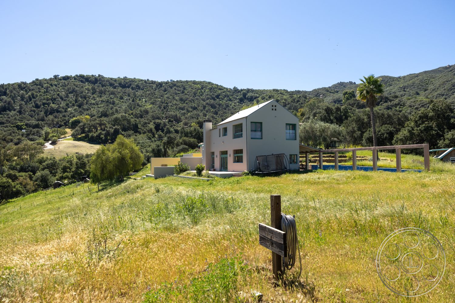 Casa del Rey - exterior, pool area, modern farmhouse style, wooden fence, sloped terrain, lush green landscape, two-story home, light-colored stucco walls, natural stone landscaping, outdoor recreational area, metal roof with angular shape, green window trim, surrounded by rolling hills, pathway leading to the house, open pasture setting, tall palm tree, broad patio area, vibrant natural light, secluded