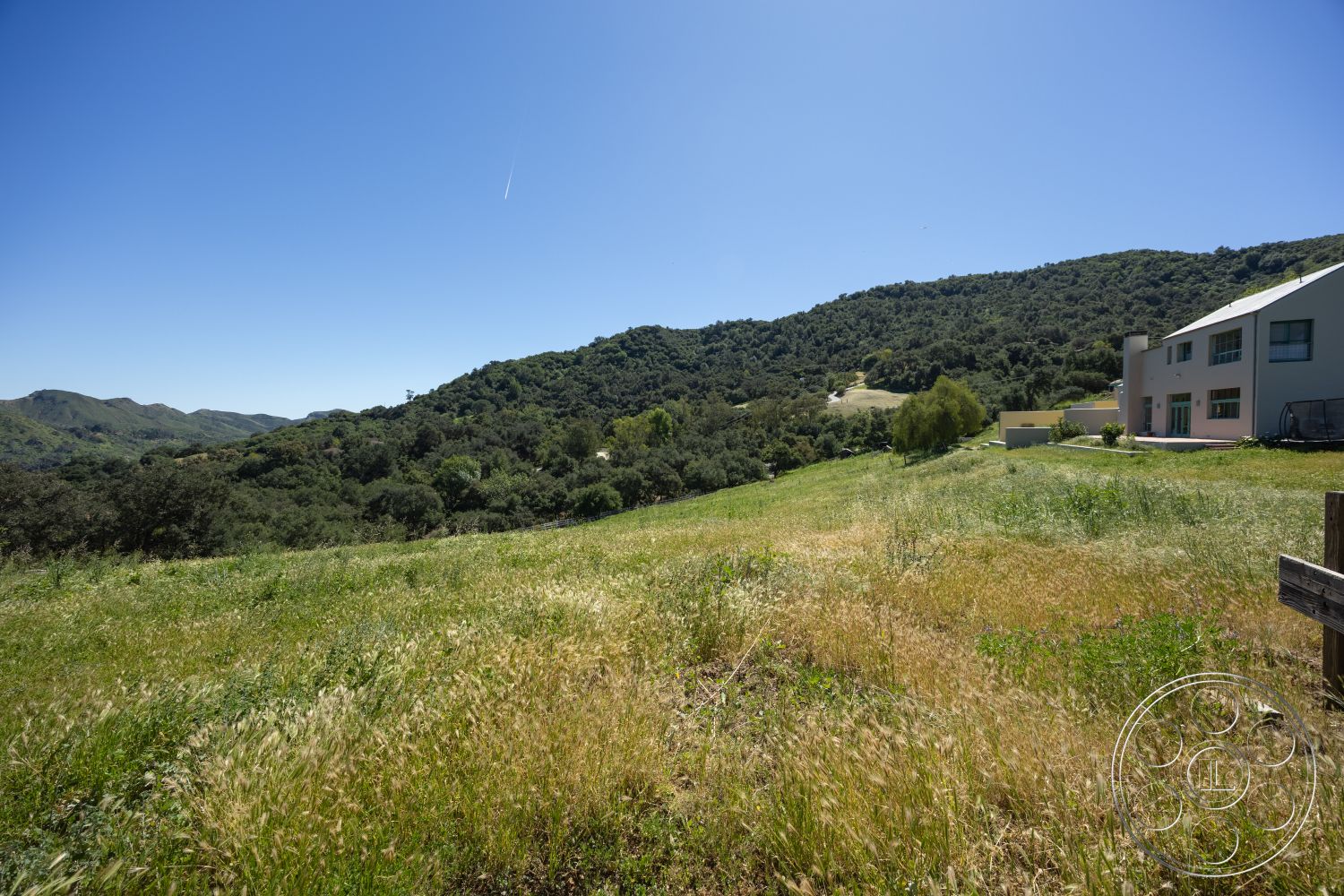 Casa del Rey - exterior, sloping terrain, minimalistic design, light-colored stucco walls, concrete driveway, mountain backdrop, large floor-to-ceiling windows, modern two-story home, concrete patio area, eco-friendly construction, rolling hillside view, natural landscape surrounding, lush greenery in foreground, urban-farmland transition, open space setting, western light exposure, sunny climate, suburban countryside