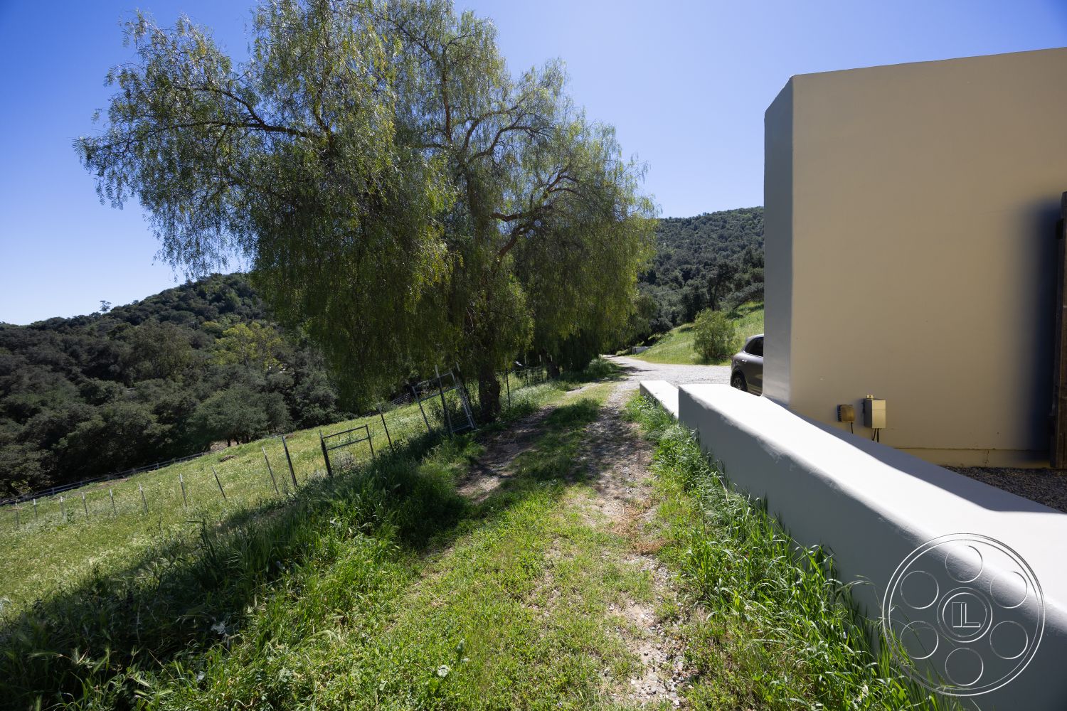 Casa del Rey - sloped terrain, natural landscape setting, sunny hillside location, smooth stucco exterior finish, exterior gravel driveway, concrete border walls, mountain view surroundings, lush green grass pathway, deciduous tree landscaping, rural countryside estate, fenced pasture area, moderate incline driveway