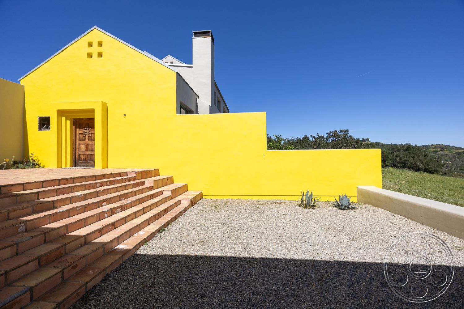 Casa del Rey - clear blue sky, wood front door, hillside location, rectangular stepping stones, angular modern architecture, exterior yellow stucco wall, terra cotta tile steps, landscaped gravel patio, low-maintenance xeriscape garden