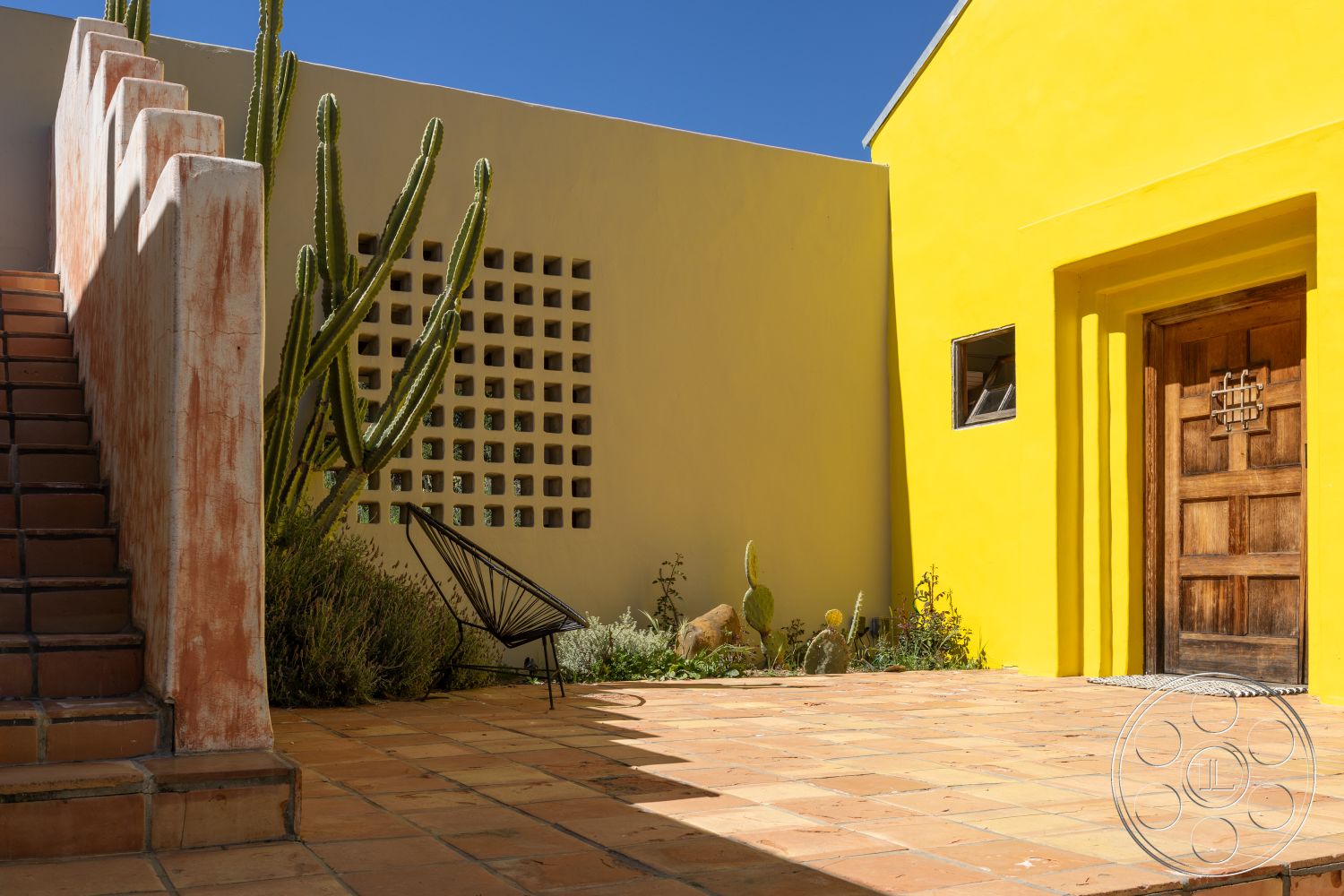 Casa del Rey - mid-century modern architectural style, exposed brick wall exterior, desert home environment, sunny courtyard setting, yellow stucco wall exterior, terra cotta tile patio flooring, sloping stairway with concrete wall, wooden front door with intricate design, cacti landscaping in courtyard setting, textured plaster finish on walls, black metal chair for outdoor seating, interior garden with native plants