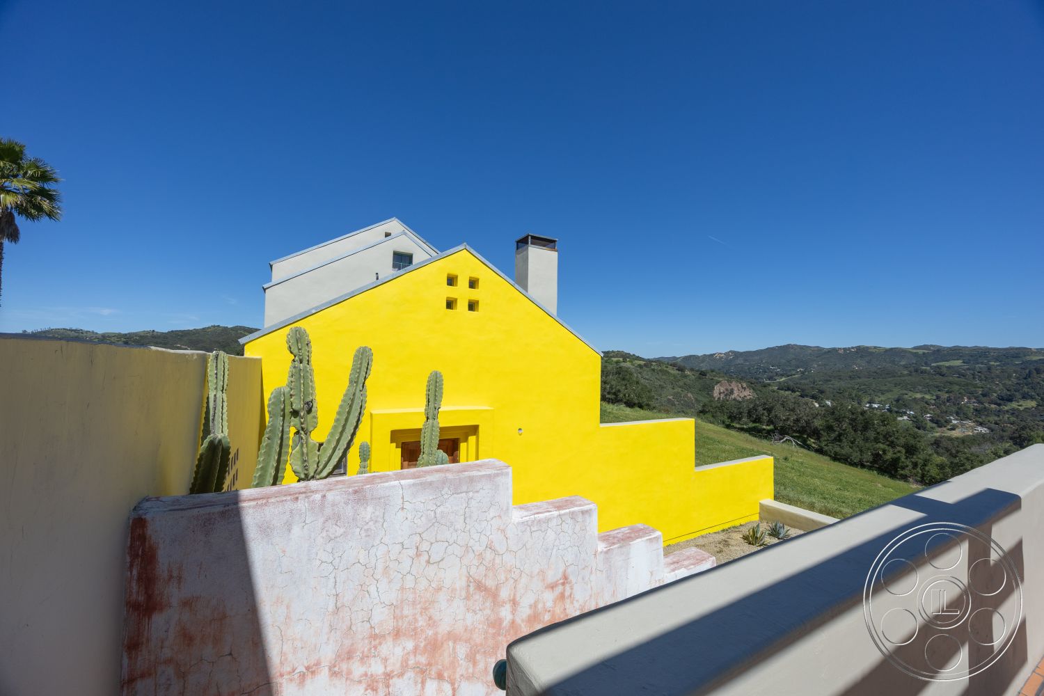 Casa del Rey - contemporary design elements, mountain view, hilltop location, minimalist architectural design, palm tree accents, modern geometric shapes, natural hillside backdrop, desert landscape setting, open-air outdoor space, concrete patio steps, cactus garden feature, exterior yellow stucco walls, terraced landscape design, angular roof lines, flat roof with parapet, rustic red plaster wall, vibrant color scheme