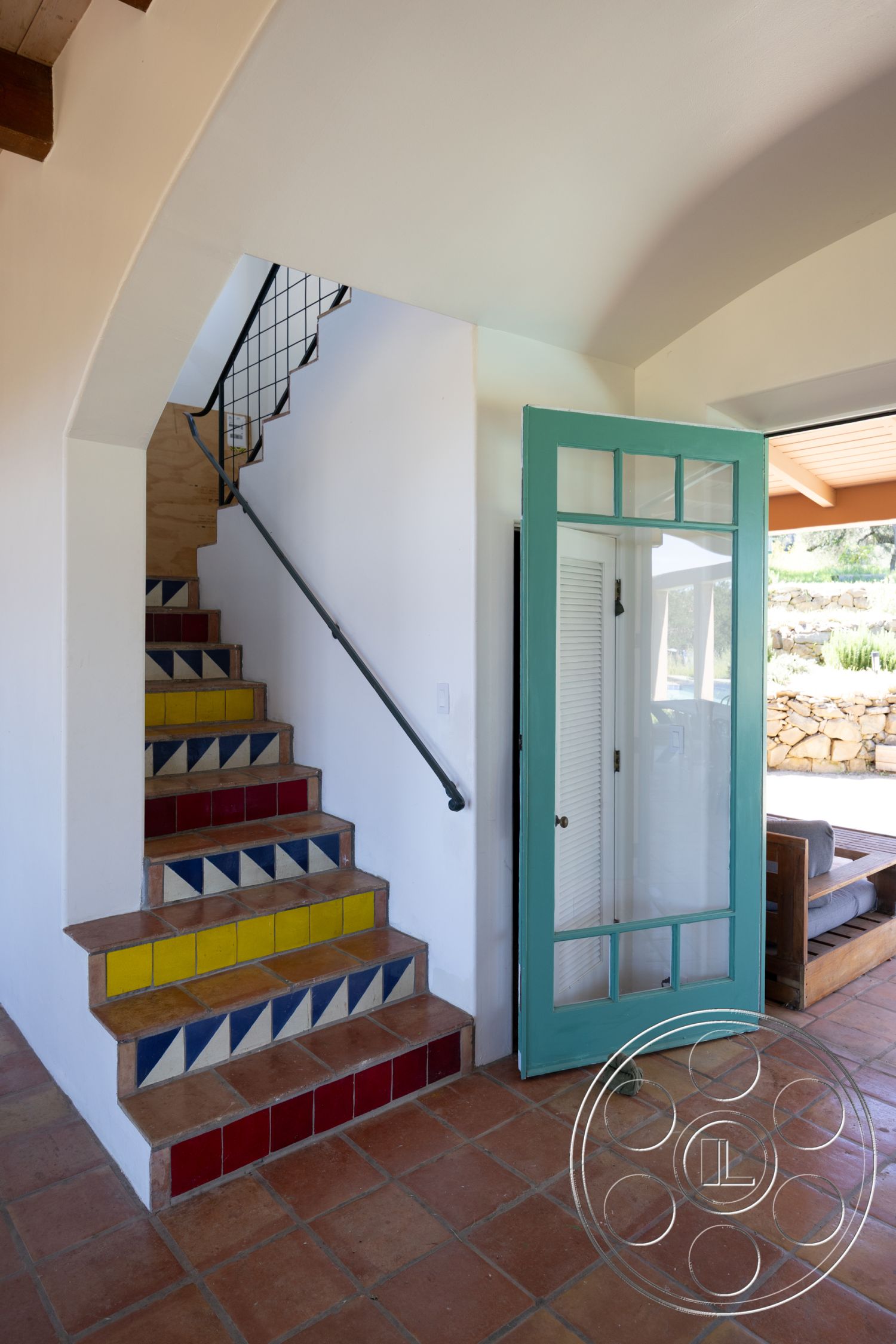 Casa del Rey - natural light, open floor plan, glass entry door, arched doorway, terracotta tile flooring, casual living space, Spanish style architecture, wood beam ceiling, metal stair railing, painted stucco walls, colorful tiled staircase risers, desert home interior, ground-level entry