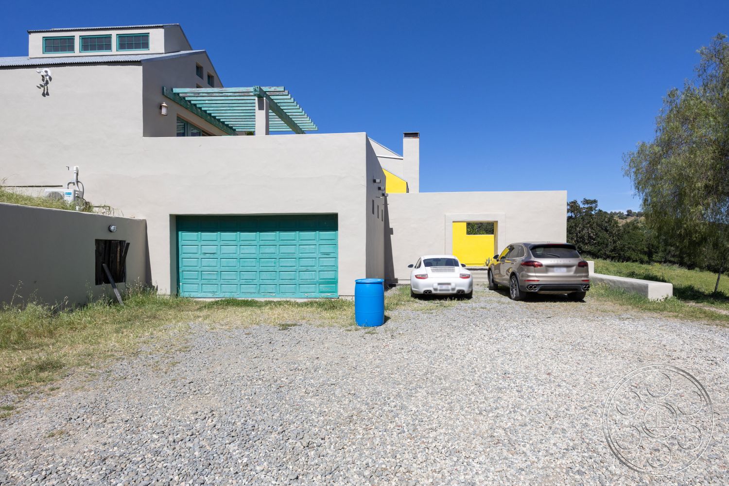 Casa del Rey - large windows for natural light, sloped terrain, modern architectural style, gravel driveway, sustainable building materials, flat roof design, exterior plaster walls, minimalist design features, rectangular house shape, turquoise garage door, green pergola structure, yellow accent walls, landscape with grassy surroundings, urban rural contrast, dual car parking area