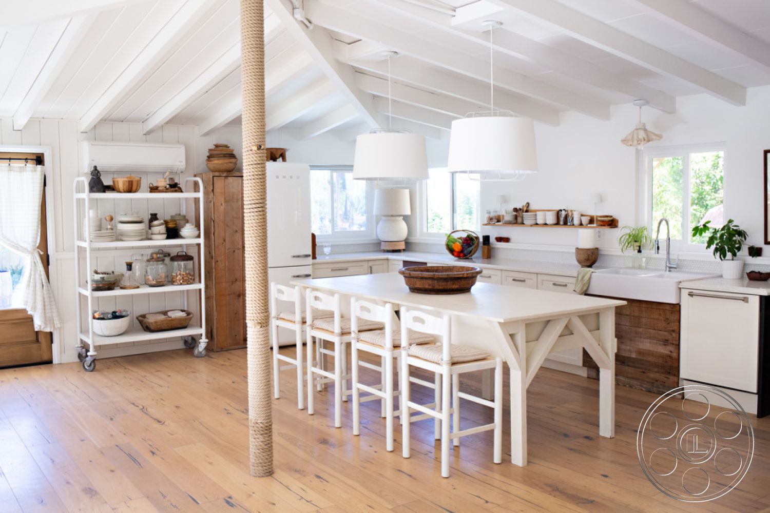 Ranch 33 - open concept layout, casual entertaining space, hardwood kitchen floors, white painted walls, shaker-style kitchen cabinets, exposed wood beam ceiling, large windows with natural light, bright and airy interior, rustic wood accents, cozy dining area, modern farmhouse kitchen, large farmhouse sink, white porcelain backsplash, isolated kitchen island, wood dining table with woven seats, minimalist décor accents, circular pendant lighting, textured wood storage cabinets, functional shelving unit on wheels, elegant fruit bowl centerpiece